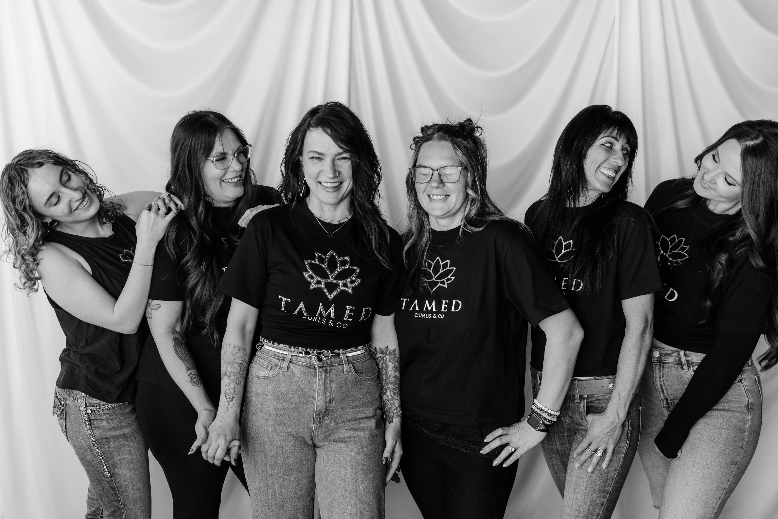 Group of six women standing together, smiling and laughing, in front of a backdrop with draped fabric. They are wearing casual clothing, some with glasses, and appear to be enjoying each other's company.