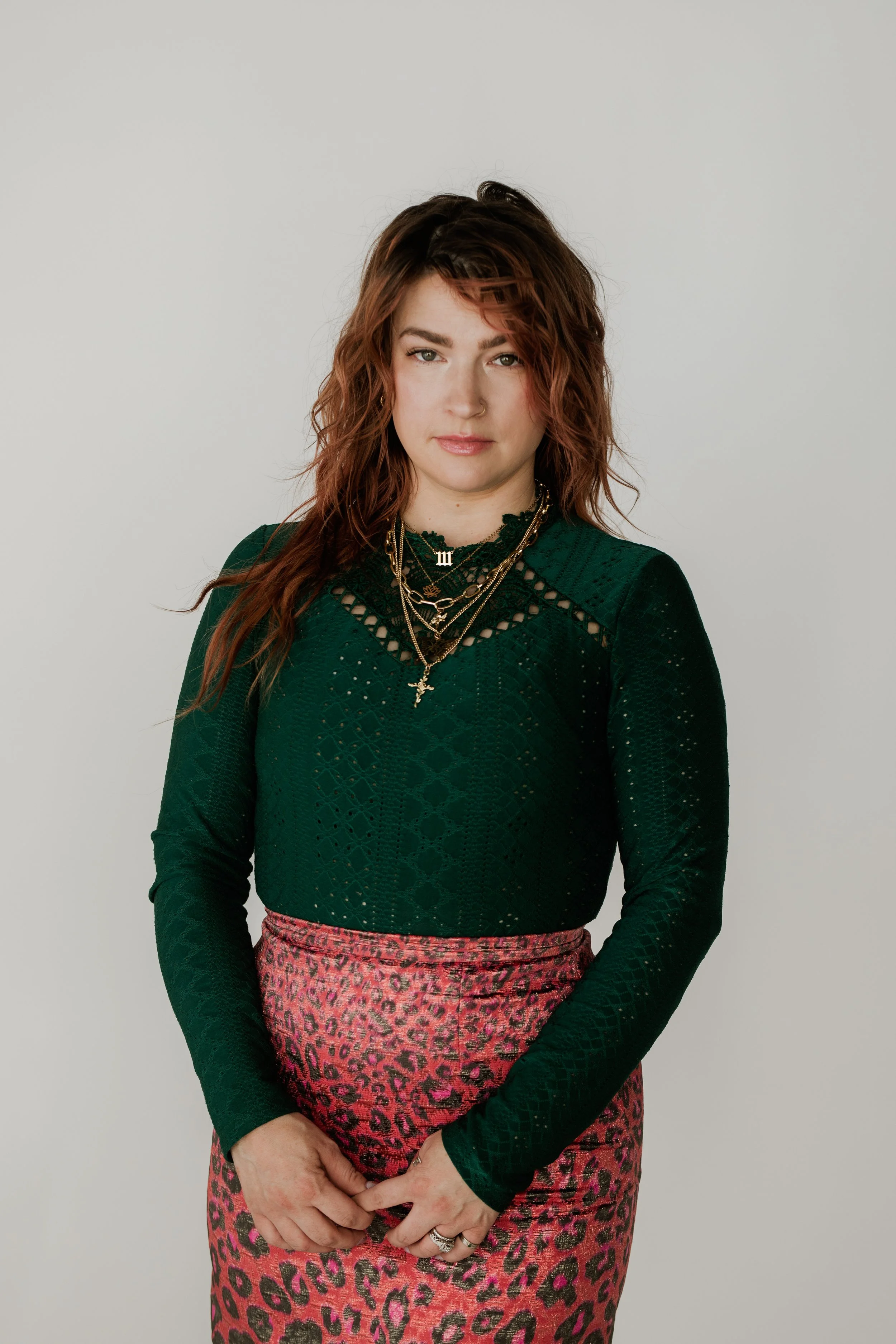 A woman with red wavy hair and light skin wearing a dark green long sleeve top with lace details, layered gold necklaces including cross and initial Charms, paired with a pink and black leopard-print skirt, stands against a plain light grey background.
