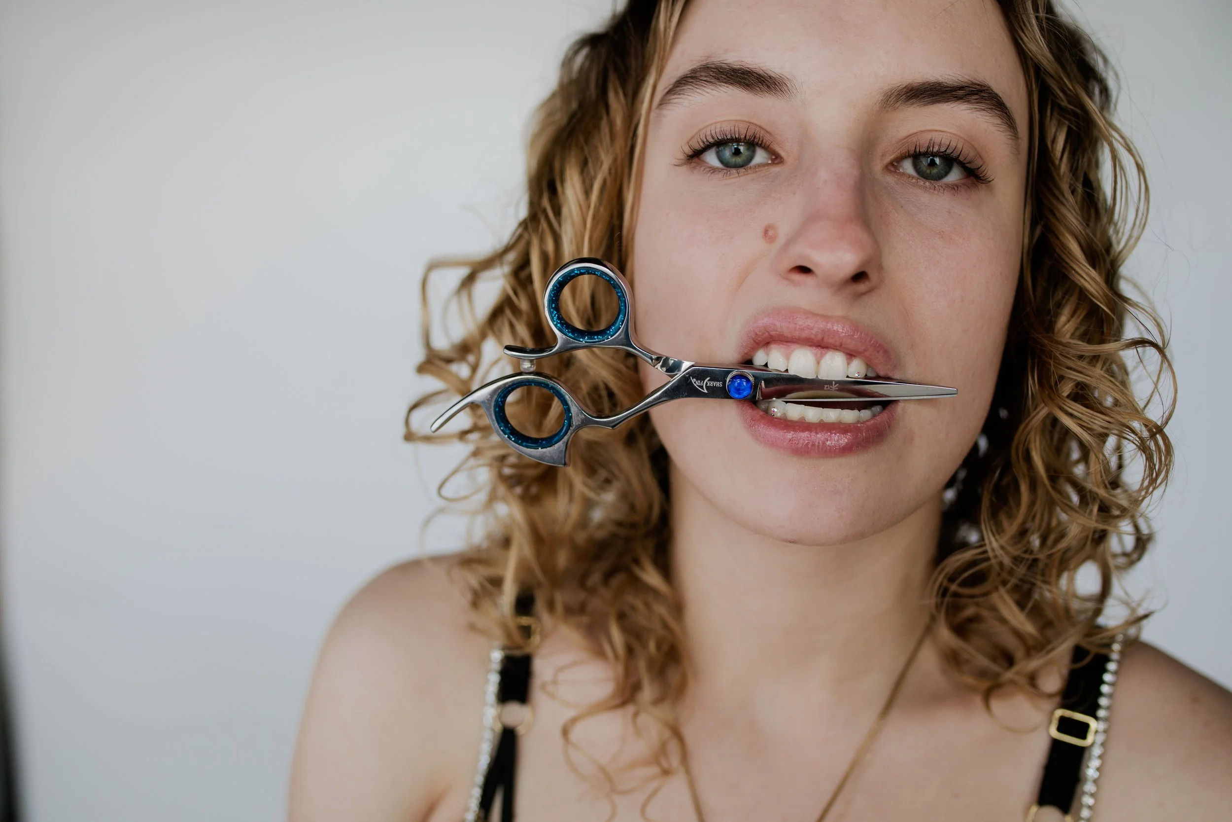 A woman with curly blonde hair and piercing blue eyes holds a pair of grooming scissors in her mouth, smiling slightly.