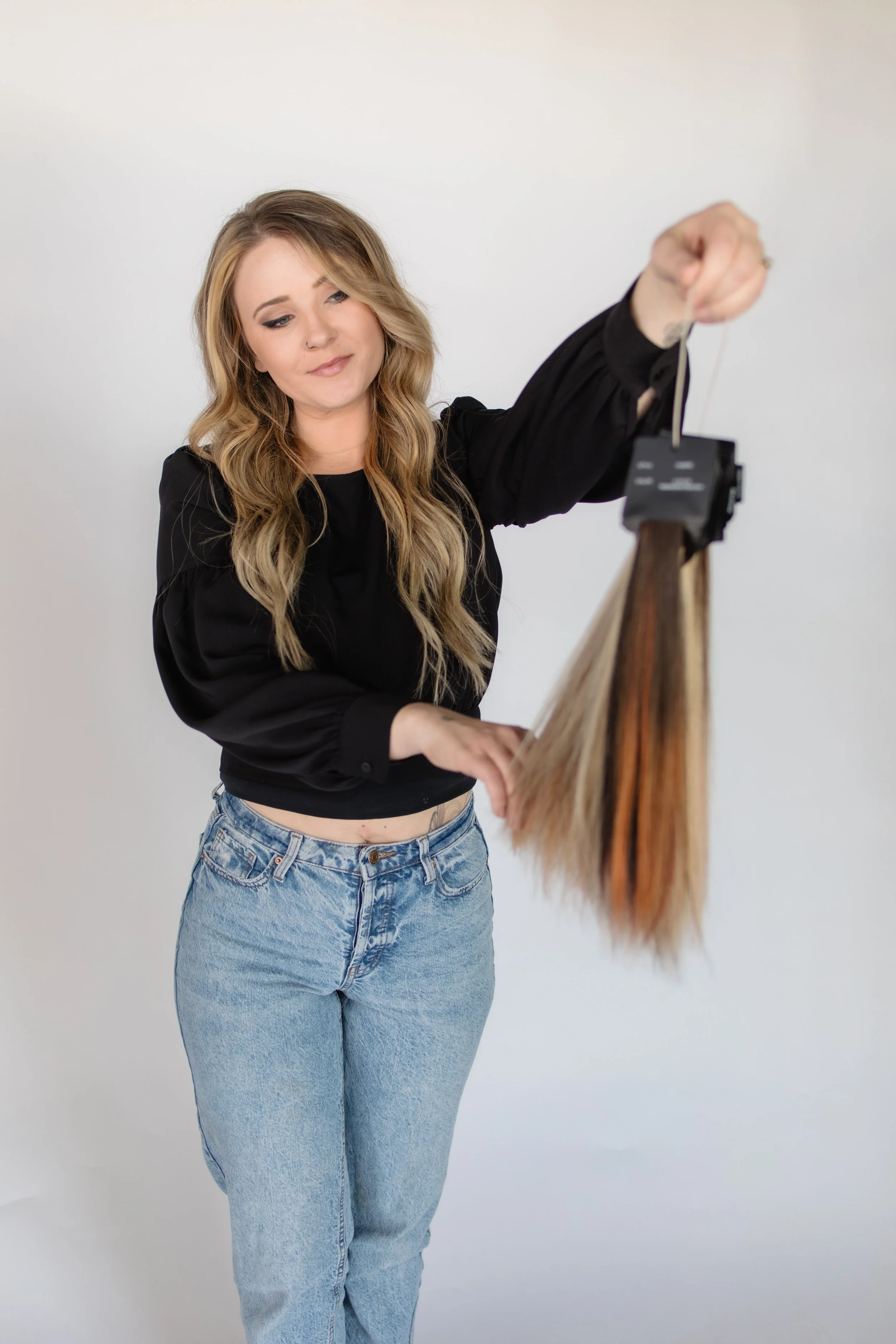 A woman with long wavy blonde hair, wearing a black blouse and high-waisted blue jeans, is smiling while holding a hanging object with long brown hair.