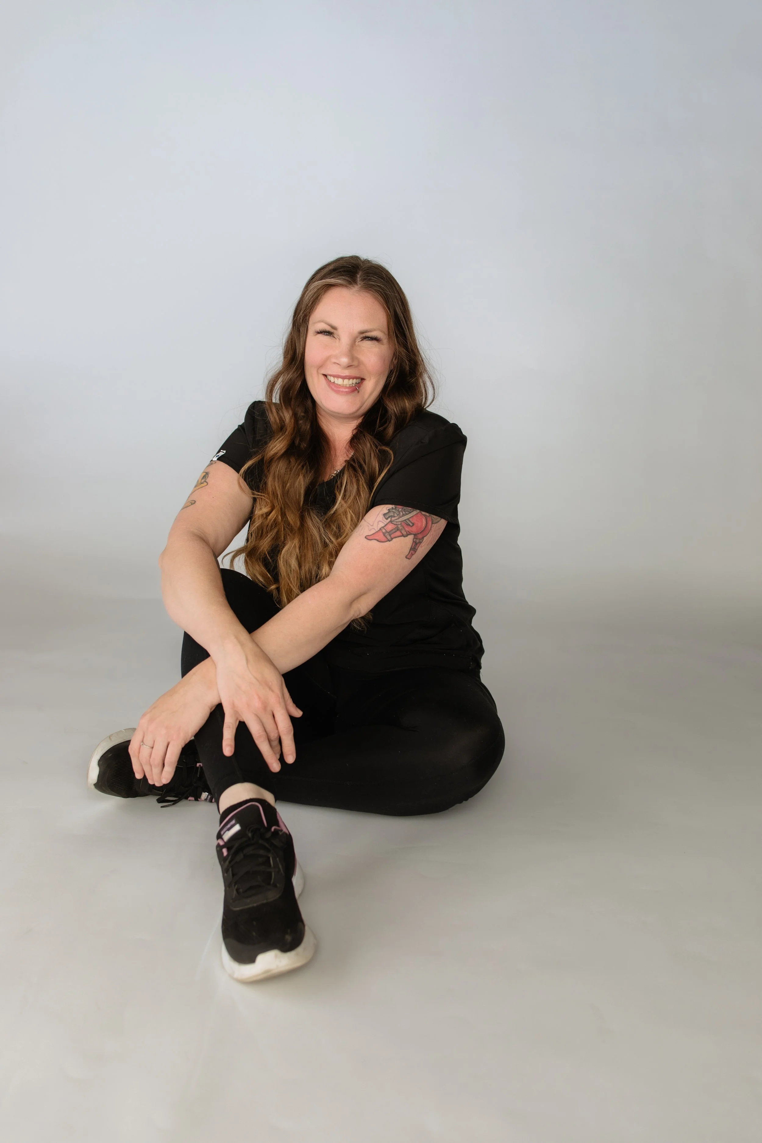 A woman with long wavy brown hair, tattoos on her arms, wearing a black t-shirt and black pants, is sitting on the floor against a plain light gray background. She is smiling, showing her teeth, and looking directly at the camera with her legs crossed and arms resting on her knees.