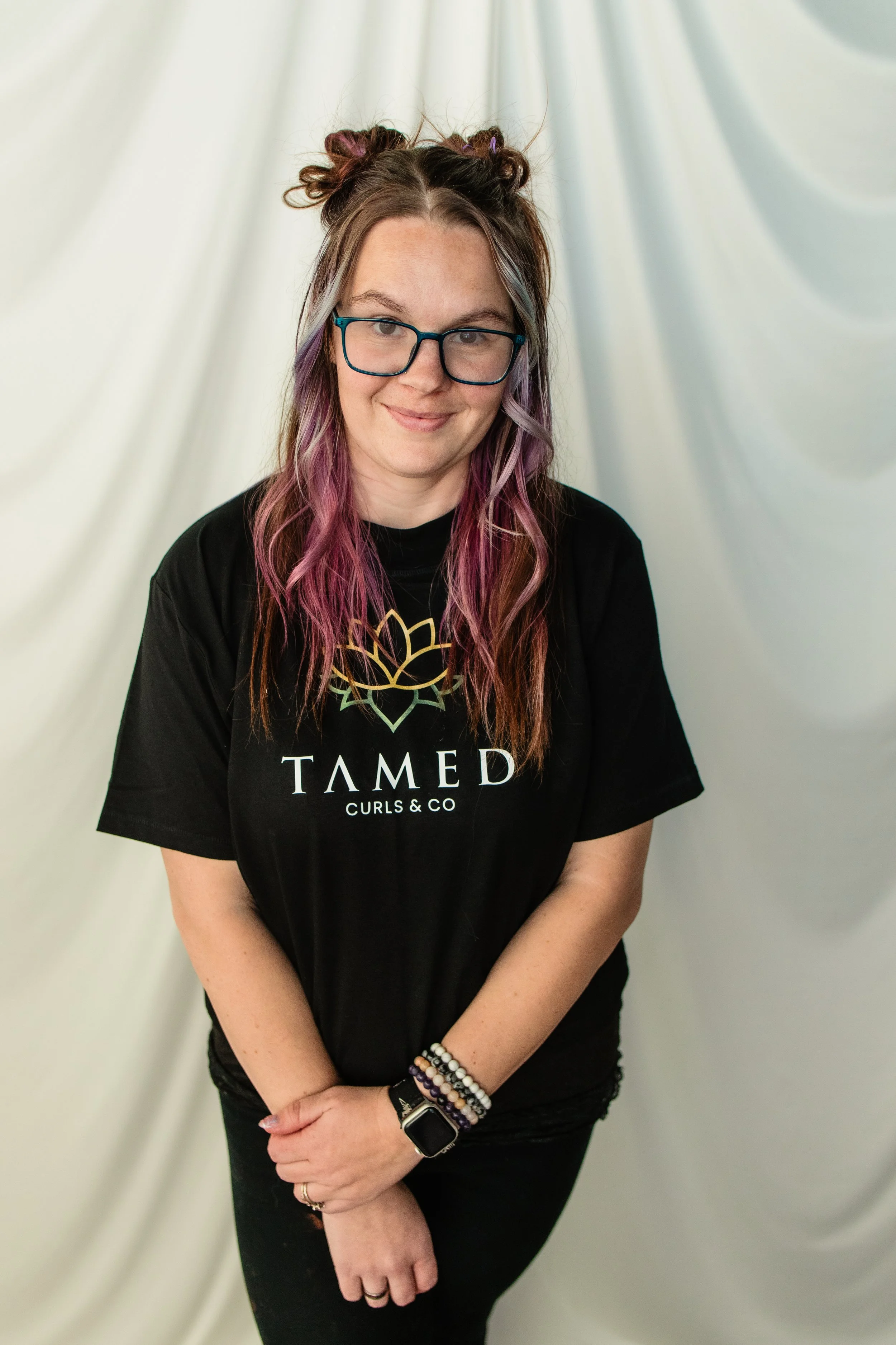 Person with purple and pink hair, wearing glasses, a black Tamed Curls & Co T-shirt, black pants, and multiple bracelets, standing in front of a white backdrop.