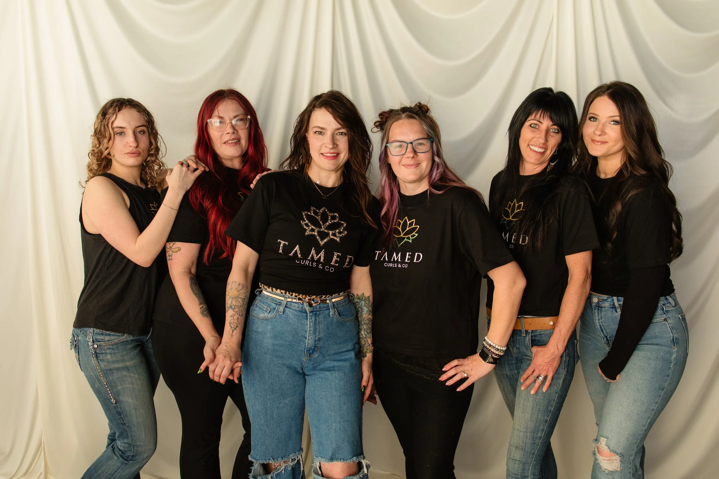 Group of six women standing together in front of a cream-colored curtain backdrop, smiling and looking at the camera. They are wearing black T-shirts with a logo that says 'TAMED' and have various hairstyles and colors, some with glasses and tattoos.