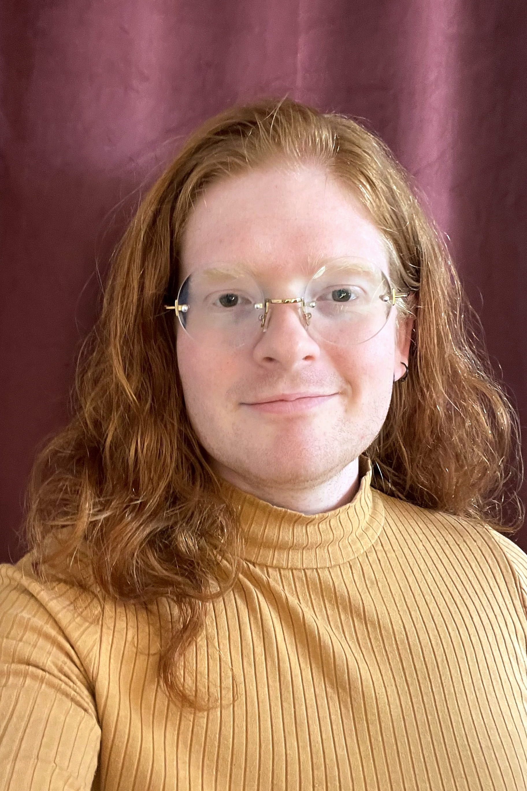 A headshot of Milo, a trans man with red hair, wearing glasses and a mustard-colored ribbed sweater, standing in front of a maroon curtain.
