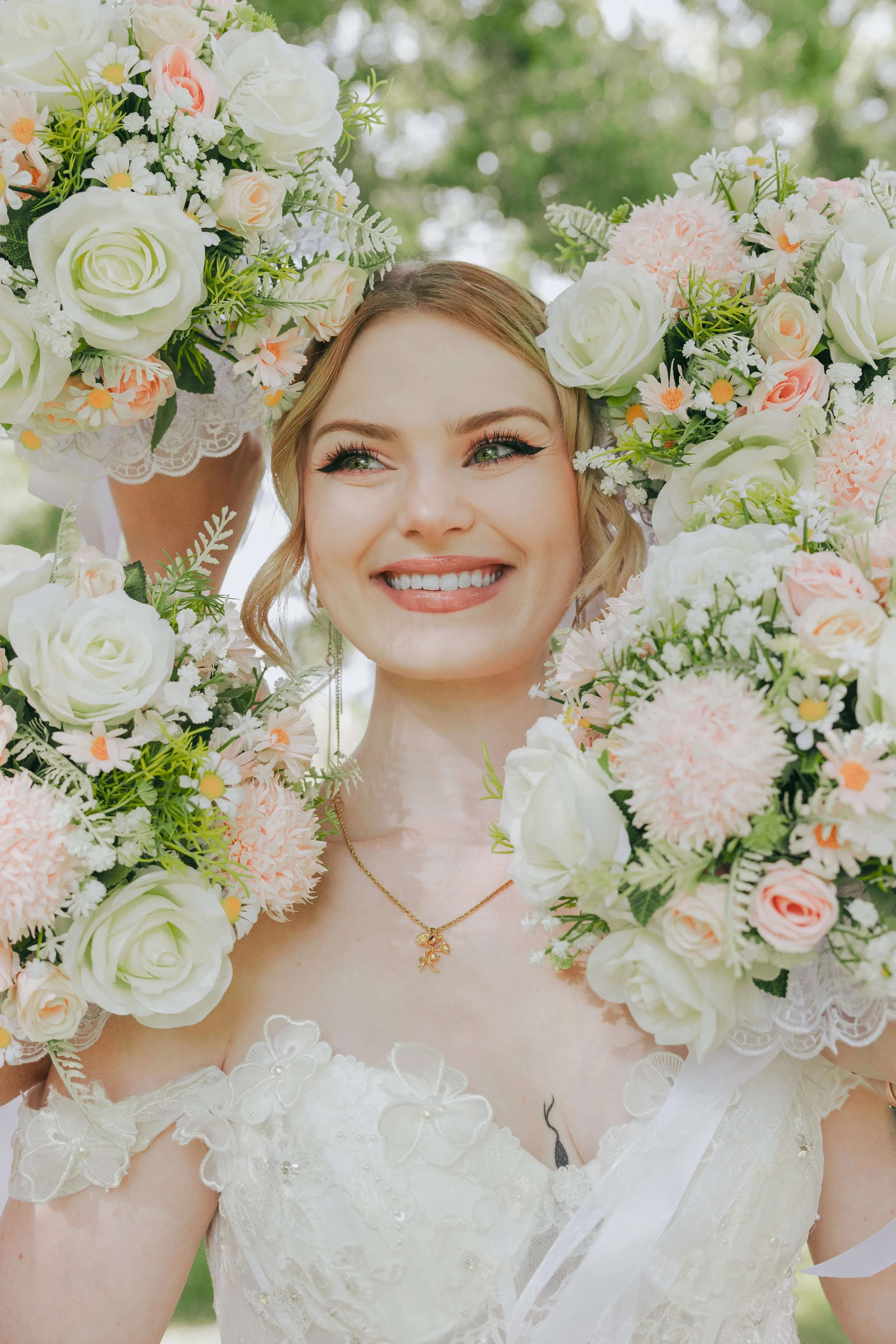 Close-up of a smiling bride wearing a floral crown and a white lace wedding dress, outdoors with green trees in the background.