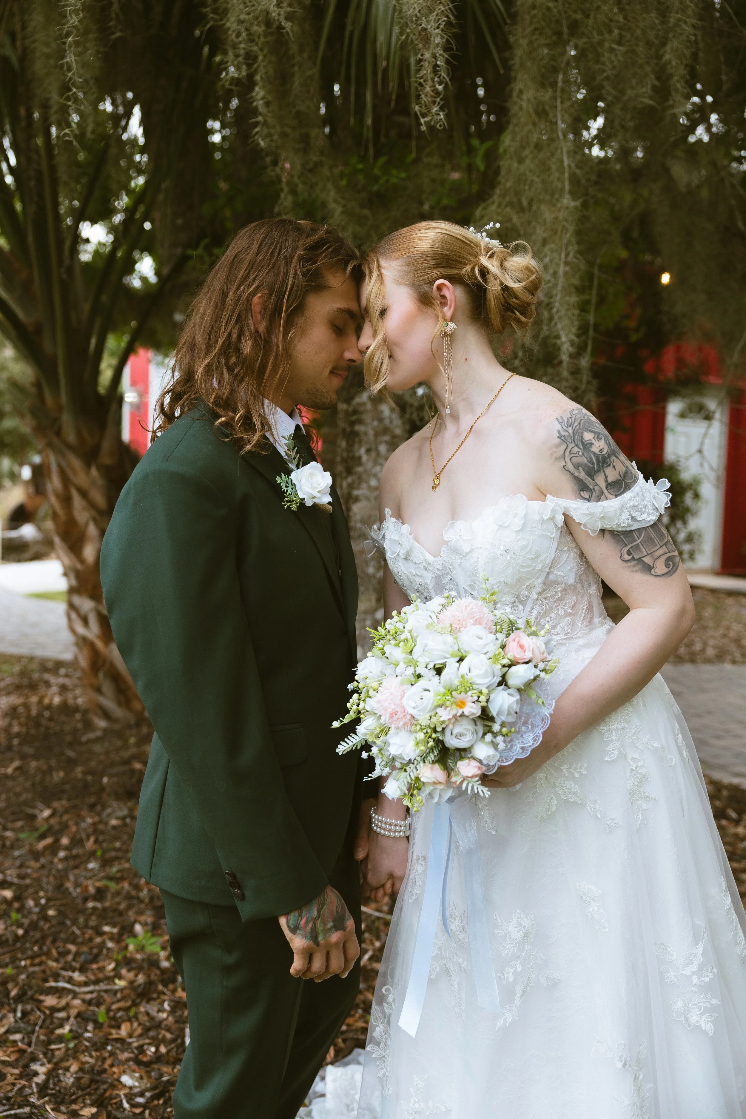 A couple is standing close with their foreheads touching during a wedding ceremony outdoors. The groom is wearing a dark suit with a white shirt and a boutonniere, and the bride is in a white lace wedding dress, holding a bouquet of white and pink flowers. They are surrounded by trees with hanging moss.