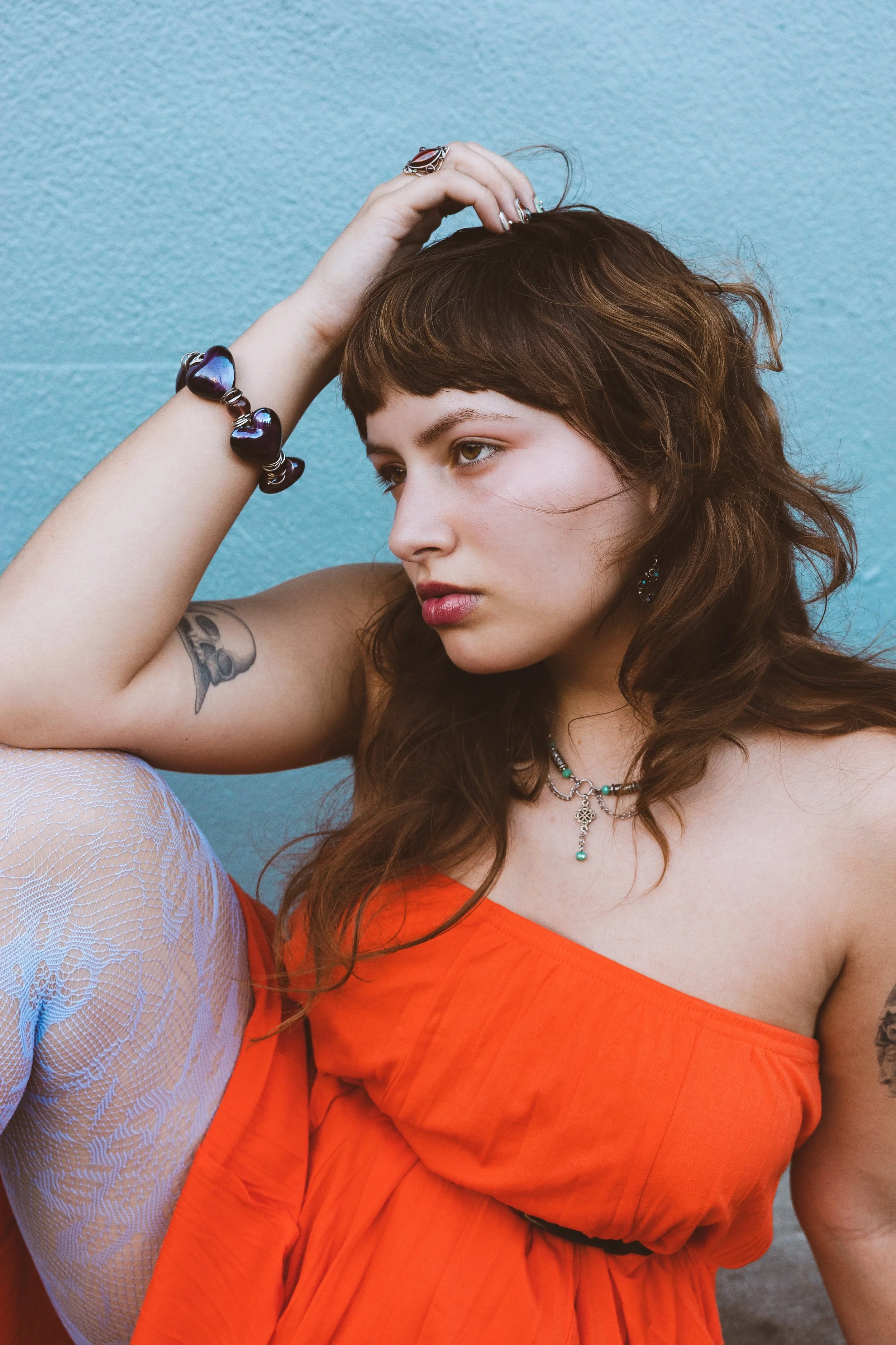 A young woman with brown wavy hair, wearing a bright orange sleeveless dress, a necklace, and earrings, posing against a light turquoise wall. She has her left arm raised, with her hand resting on her head, showcasing a large ring and a purple bracelet.
