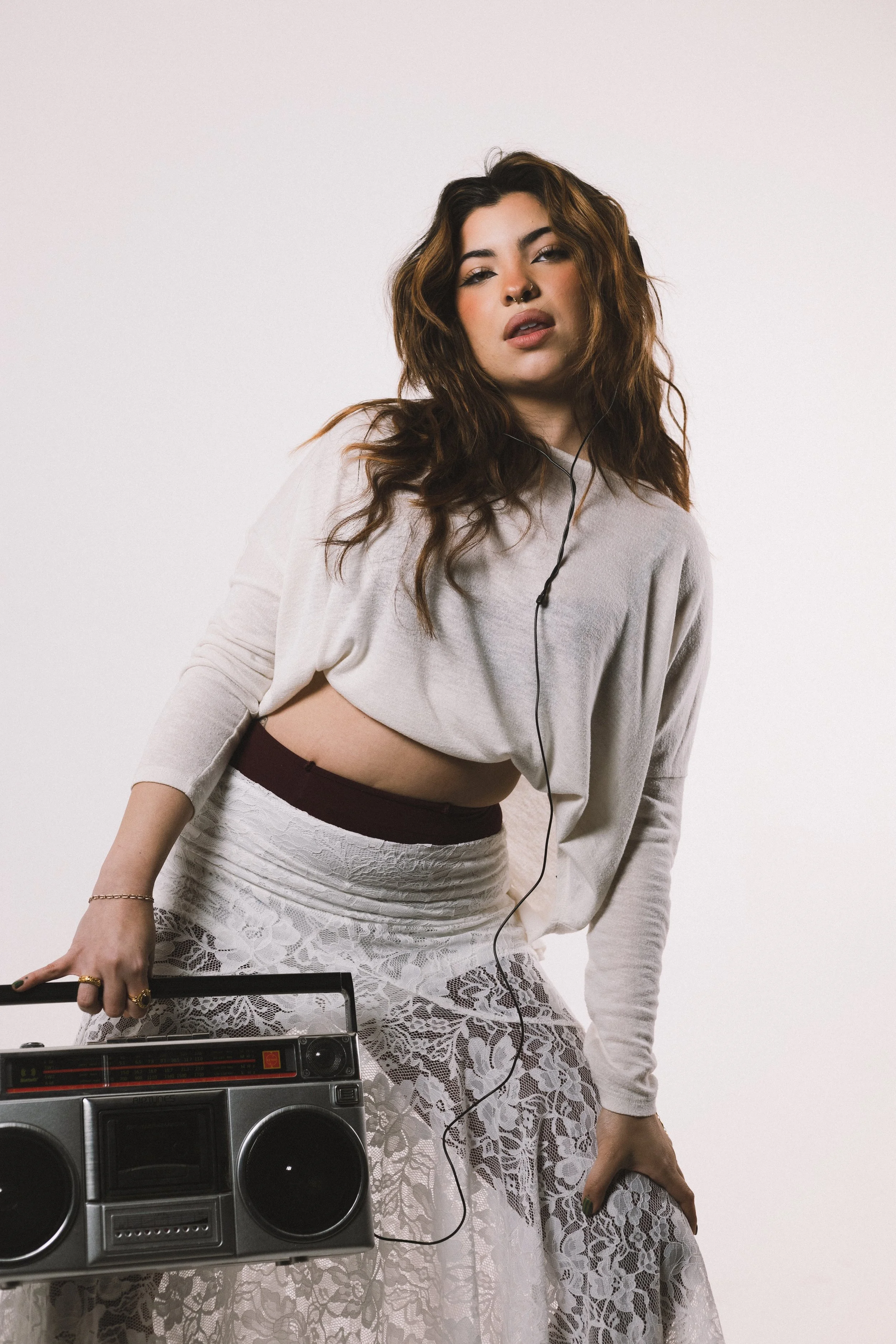 A young woman with wavy brown hair, wearing a beige crop top, lace skirt, and multiple rings, stands next to a vintage boombox, holding a microphone, against a plain white background, with earbuds in her ears.