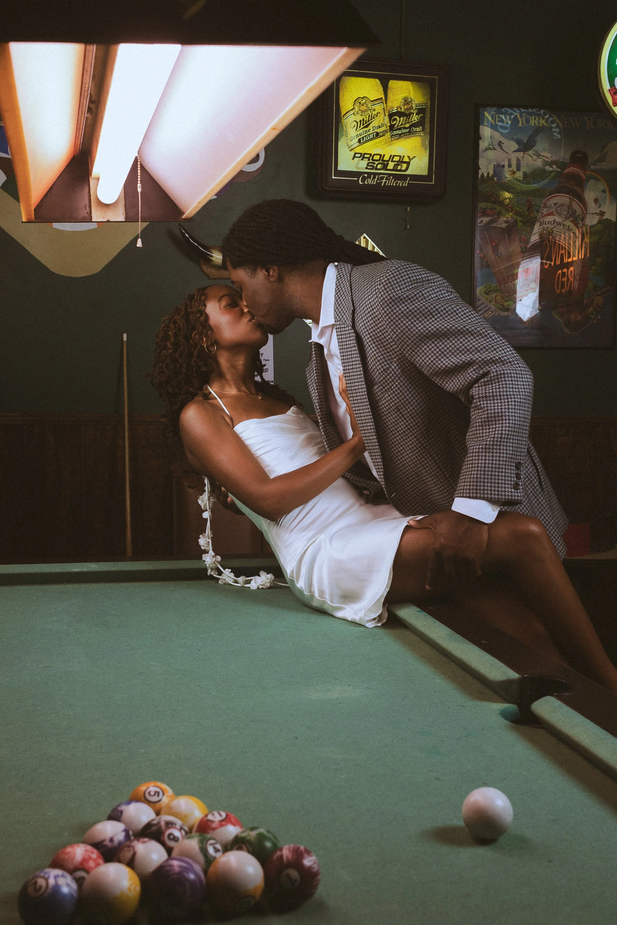 A couple in formal attire kissing over a pool table in a bar with vintage signs on the wall, including Miller beer and New York posters, with a ball rack at the edge of the pool table and a cue ball near the center.