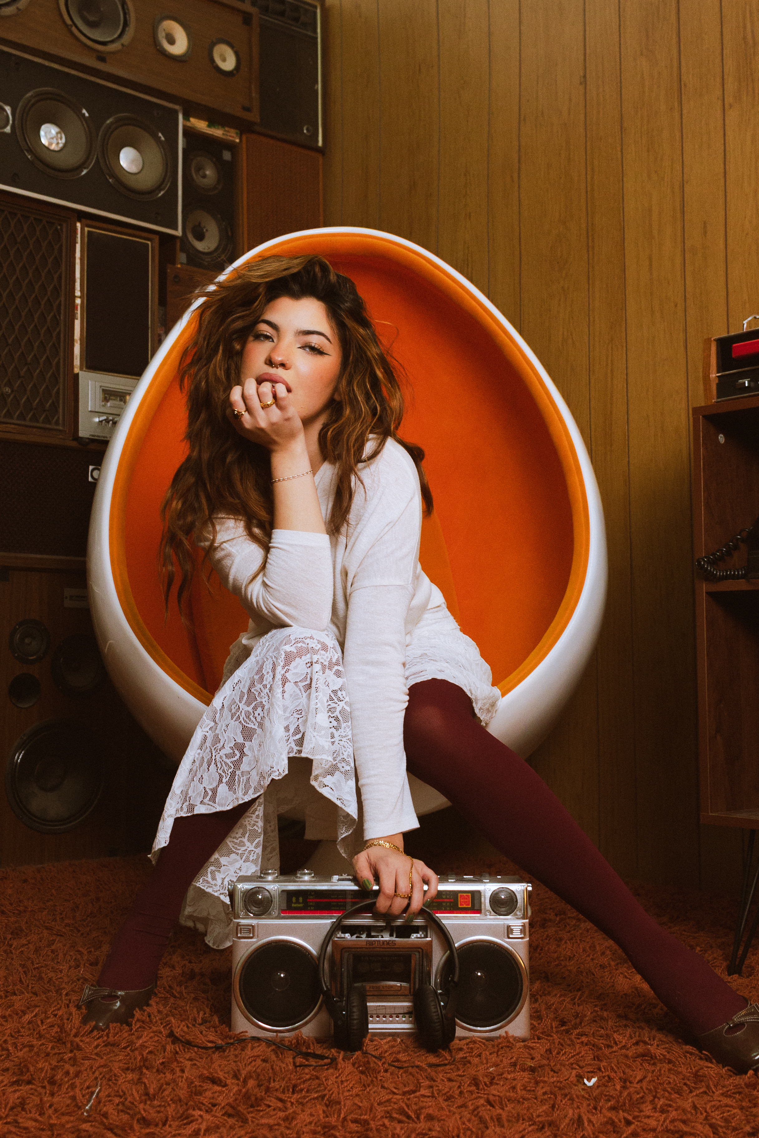 A young woman sitting in an orange circular chair, dressed in a white lacy dress and maroon tights, with wavy brown hair, surrounded by vintage speakers and a boombox in a retro room.