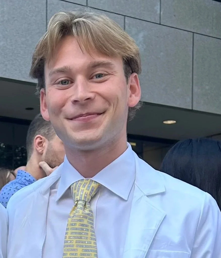 A young man with light brown hair and blue eyes smiling, wearing a white shirt and a yellow tie with a patterned design, standing outdoors near a modern building.