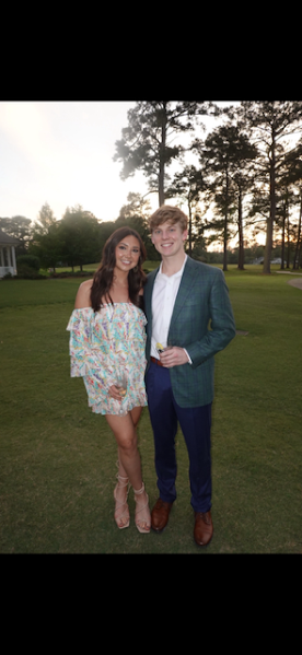 A woman and a young man standing outdoors on a grassy area with trees and sunset in the background. The woman is wearing a colorful off-the-shoulder dress and beige high heels. The young man is wearing a green checkered blazer, white shirt, blue pants, and brown shoes, holding a drink.