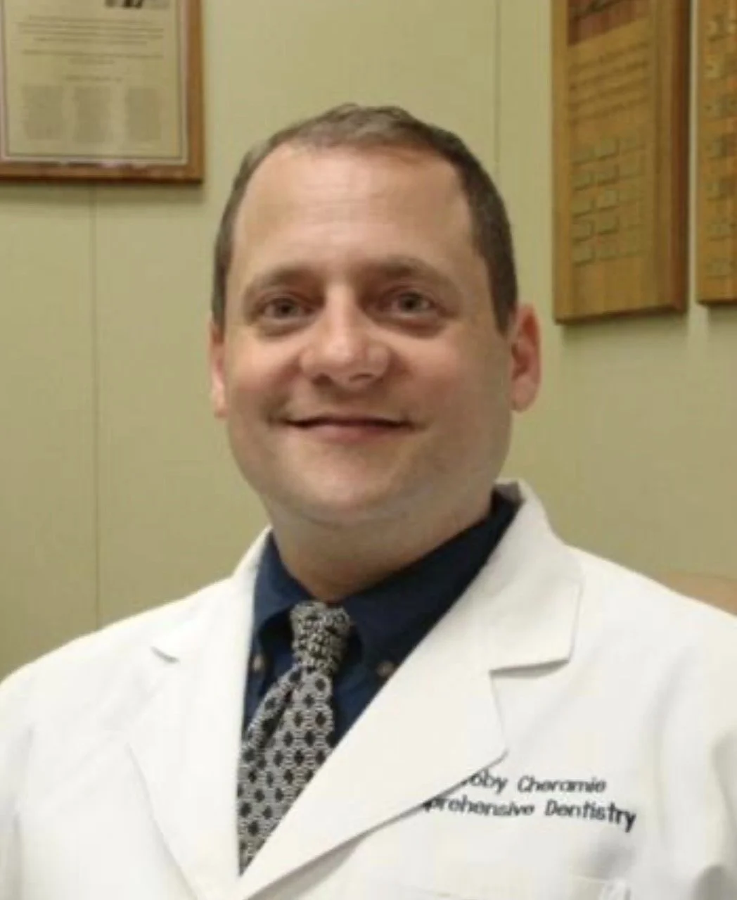 A man wearing a white dental coat, with a name patch that reads "Baby Chavarro, Orthodontic Dentistry". He is smiling and standing indoors, with framed posters on the wall behind him.