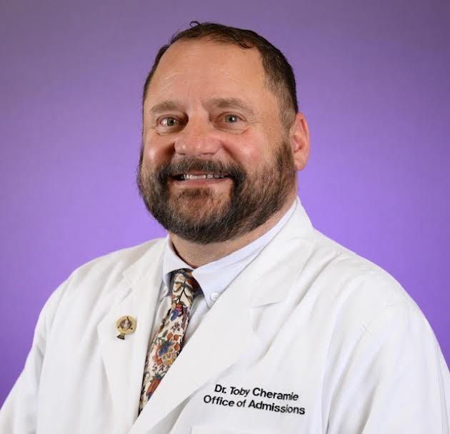 A man with a beard and brown hair, wearing a white medical coat and a patterned tie, smiling at the camera against a purple background.