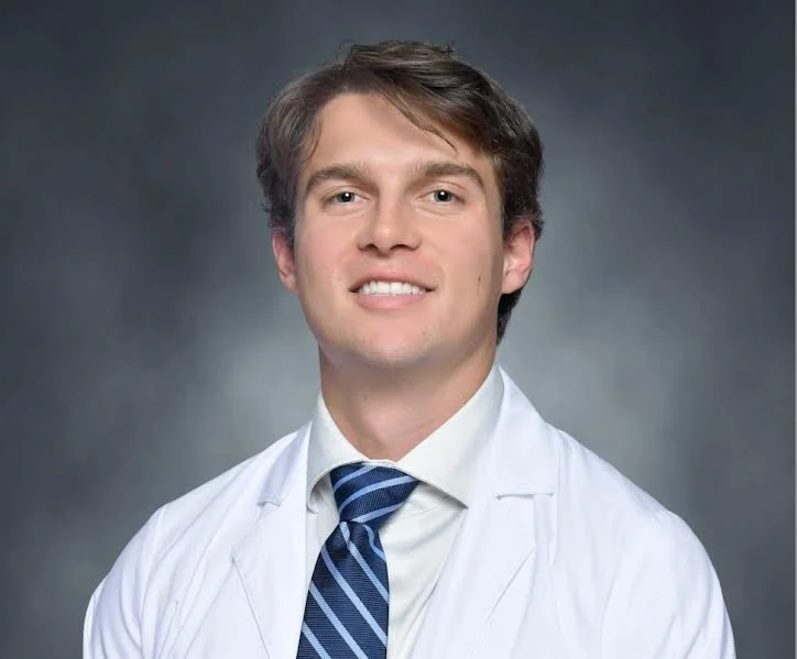 Portrait of a young male doctor with brown hair, wearing a white coat, white shirt, and blue striped tie, against a dark gray background.