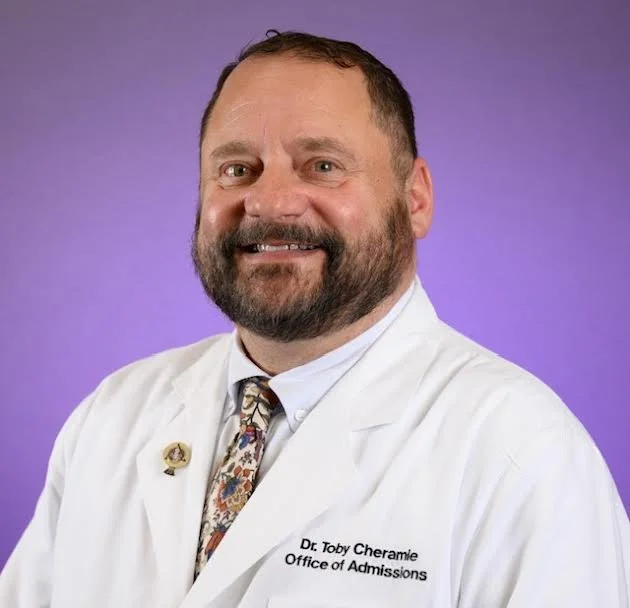 Professional portrait of a man with a beard and short hair, wearing a white lab coat with a name tag that says Dr. Toby Cheramie, Office of Admissions, and a patterned tie, against a solid purple background.
