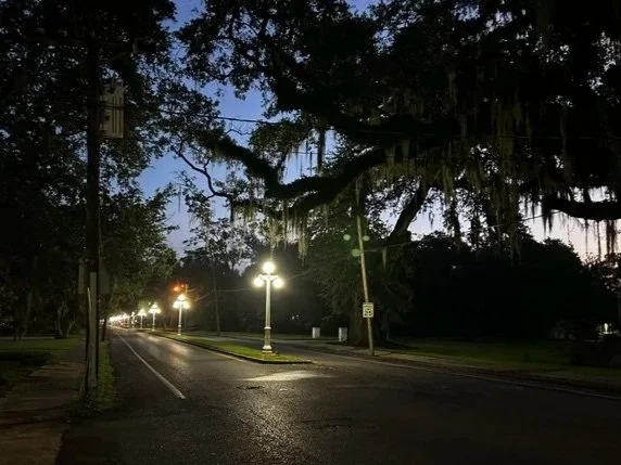 A quiet street at dusk with streetlights, trees, and a cloudy sky.