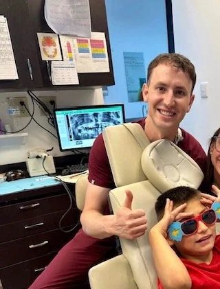 Smiling man giving a thumbs-up next to a young boy wearing sunglasses in a dentist's office.