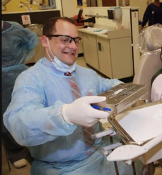 A man in medical attire, including a gown, gloves, mask, and glasses, is smiling while holding a metal tray and a blue item in a clinical or laboratory setting.