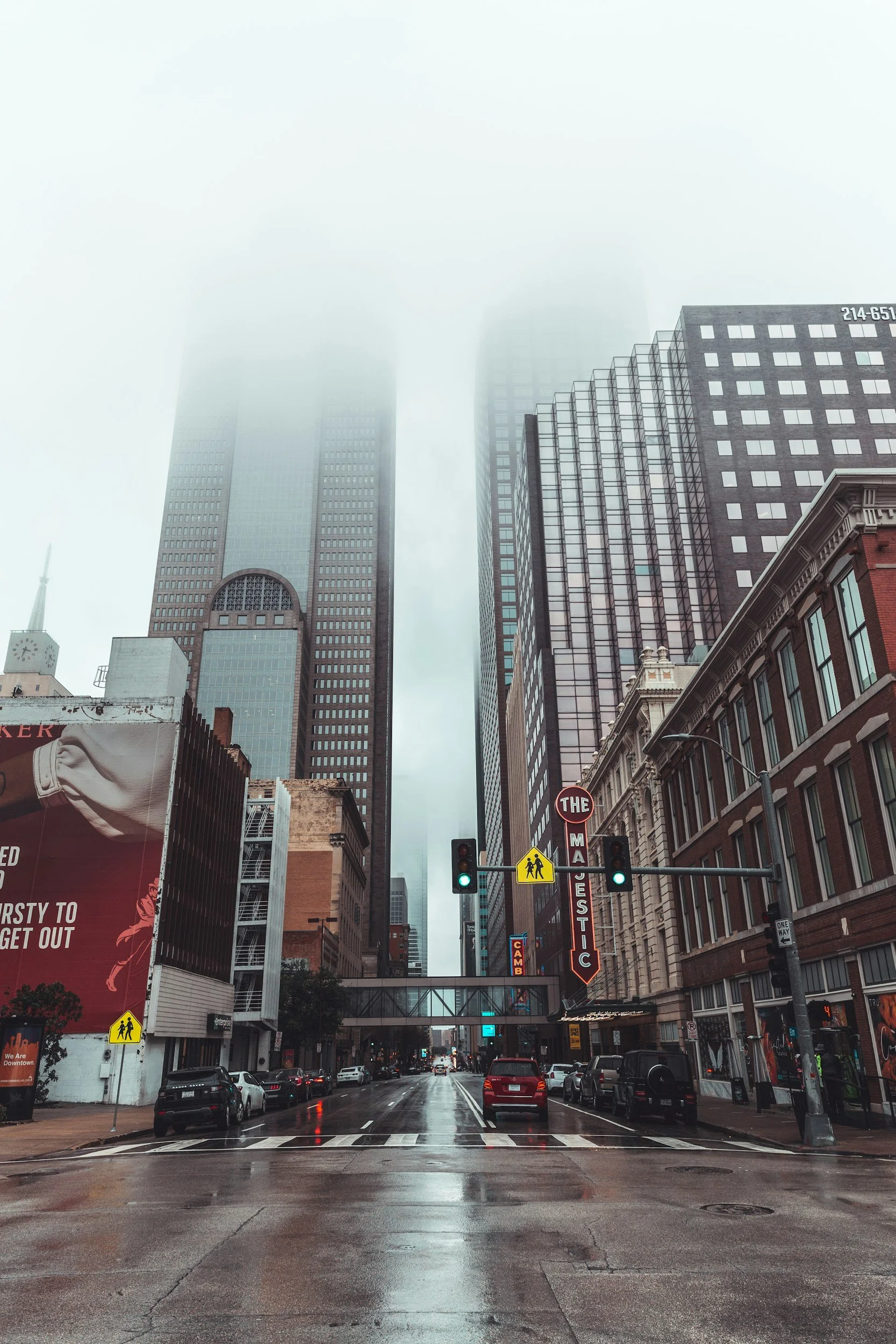 Rainy city street scene with traffic lights, cars parked along the curb, tall skyscrapers covered in fog, pedestrian crossing sign, and buildings with various signage and architecture.