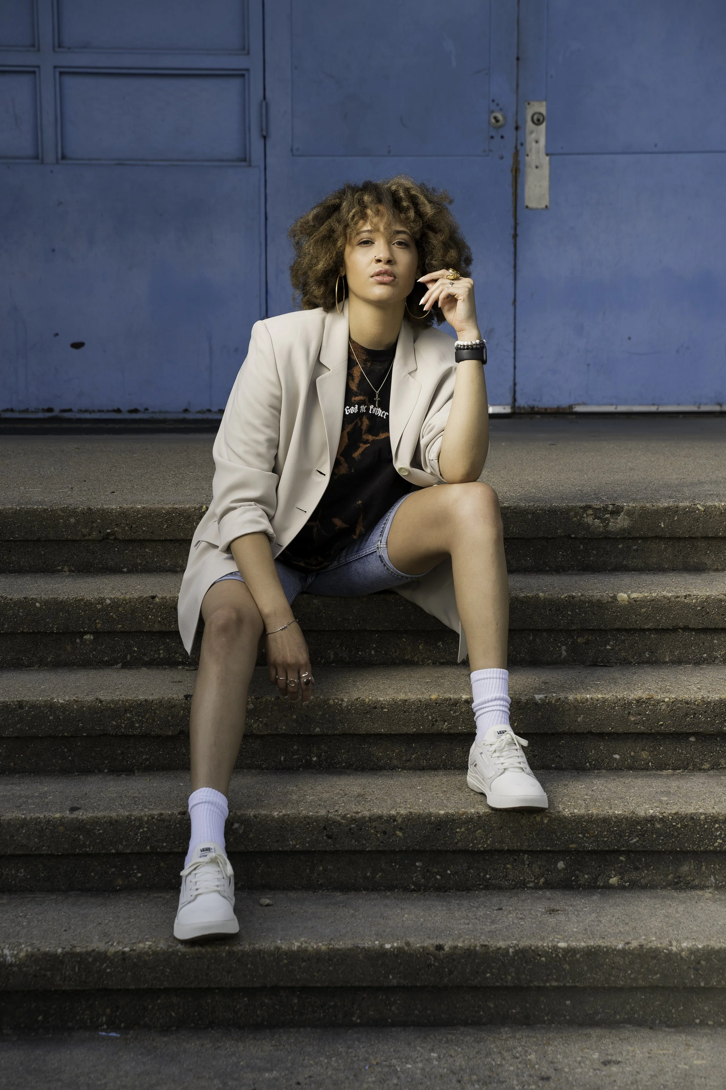 A young woman with curly hair sitting on outdoor concrete steps in front of a blue metal door. She is wearing a beige blazer, black graphic t-shirt, denim shorts, white sneakers, white socks, and jewelry including rings, hoop earrings, and necklaces.