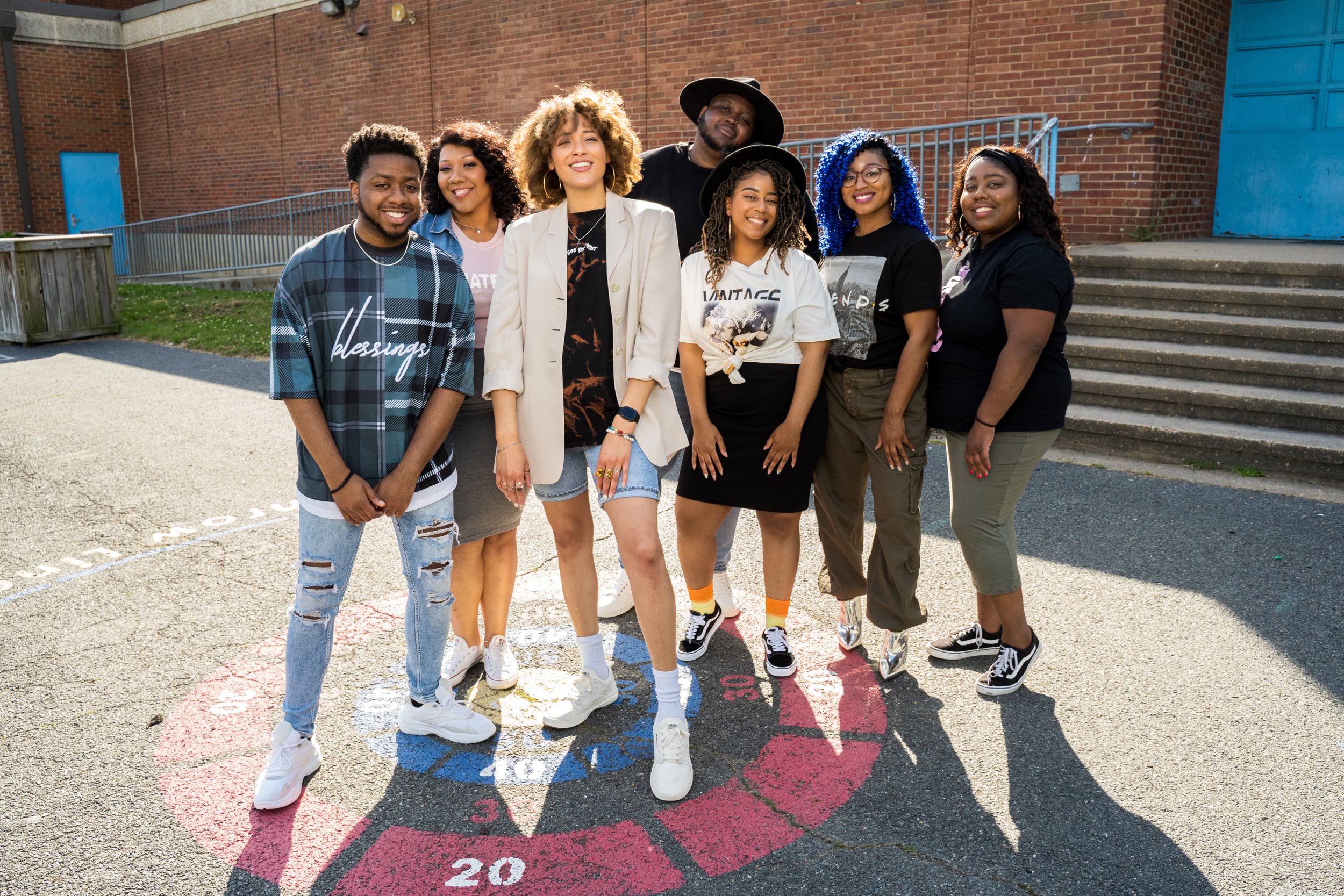 Group of seven diverse young adults standing together outdoors on a sunny day, smiling for the camera in front of a brick building with stairs.