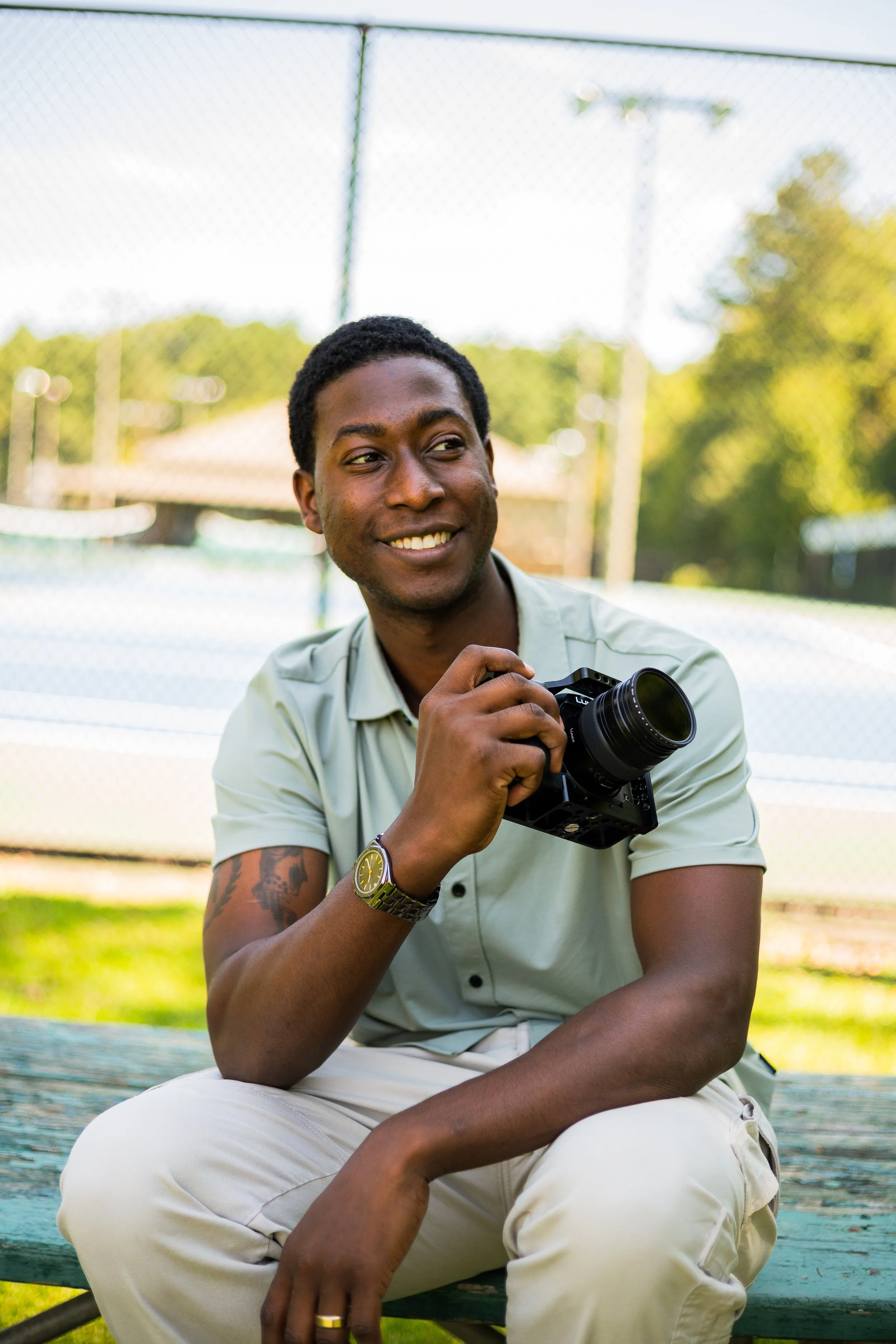 A young man sitting on a park bench outdoors, smiling and holding a camera, with a sports field and trees in the background.
