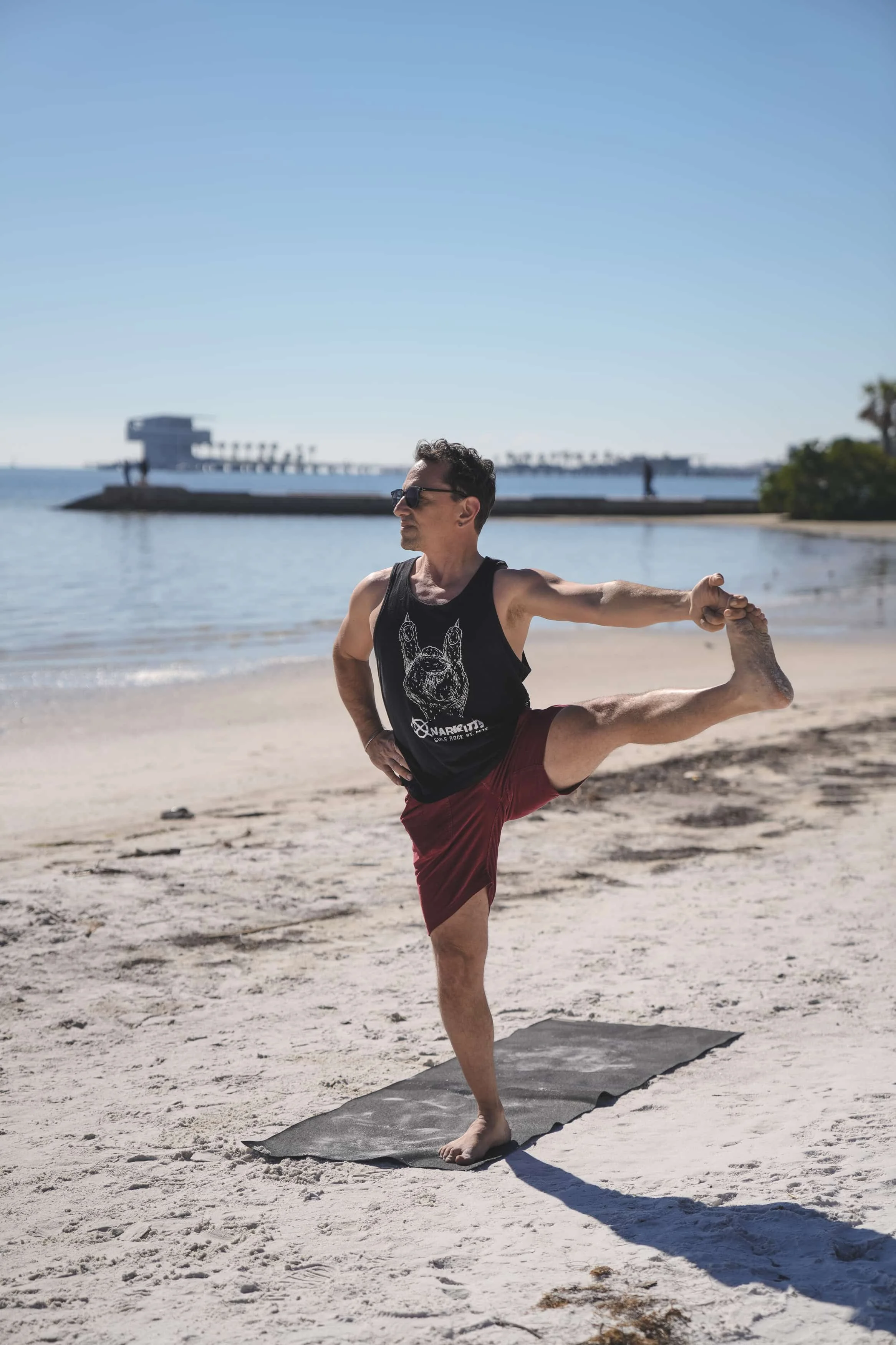 A man practicing yoga on a beach, standing on one leg in a yoga pose with a bent knee and holding his foot behind him, wearing sunglasses and a tank top with a dog design. The background features the ocean, a pier, and a clear blue sky.