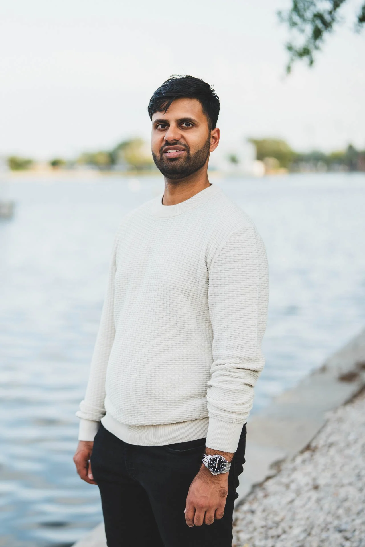A man with dark hair and a beard standing near a body of water, wearing a white textured sweater and a watch.