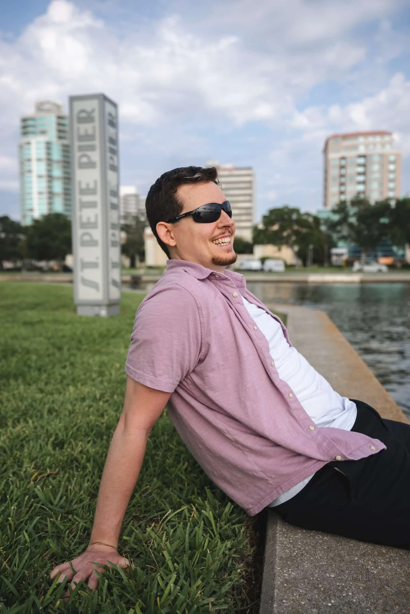 A smiling man wearing sunglasses and a pink short-sleeve button-up shirt, sitting on a concrete ledge by a river, with modern buildings and trees in the background, under a partly cloudy sky.