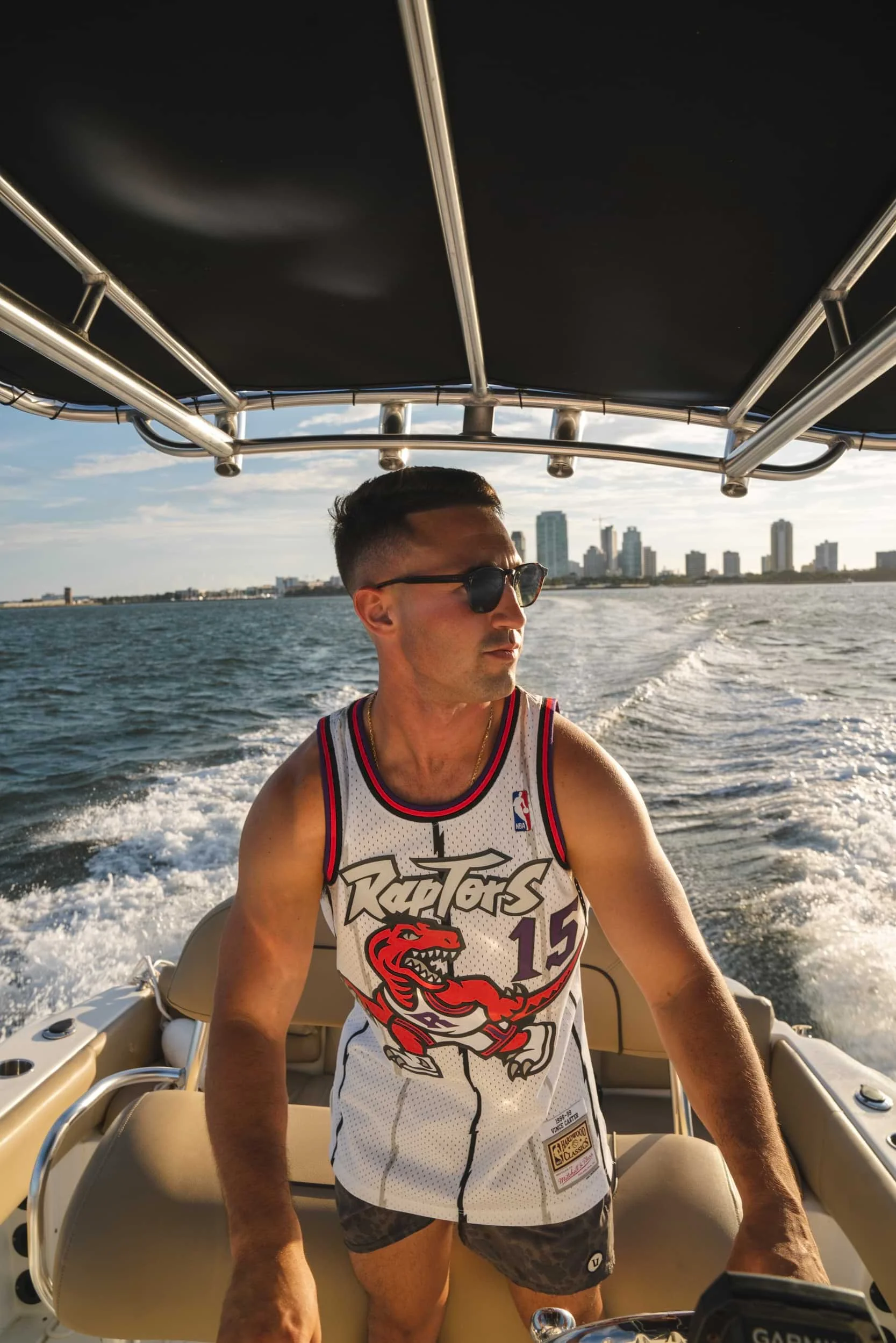 A man wearing sunglasses and a Raptors basketball jersey driving a boat on the water with the city skyline in the background.
