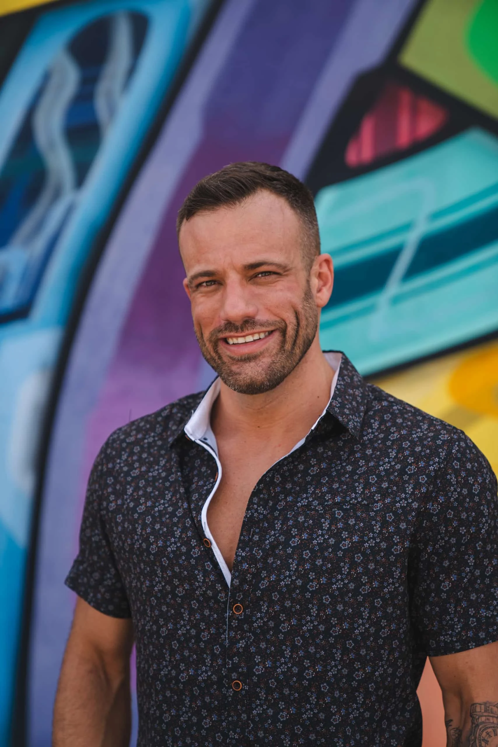 A smiling man with short dark hair and a beard, wearing a dark floral shirt with a white collar, standing in front of a colorful mural with abstract shapes and vibrant colors.