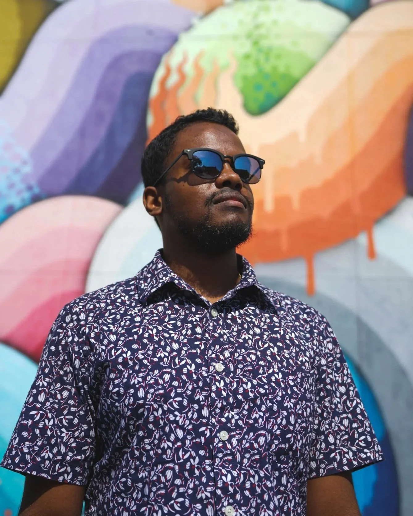 A man wearing sunglasses and a patterned shirt standing in front of a colorful mural of large, abstract, rainbow-colored shapes.