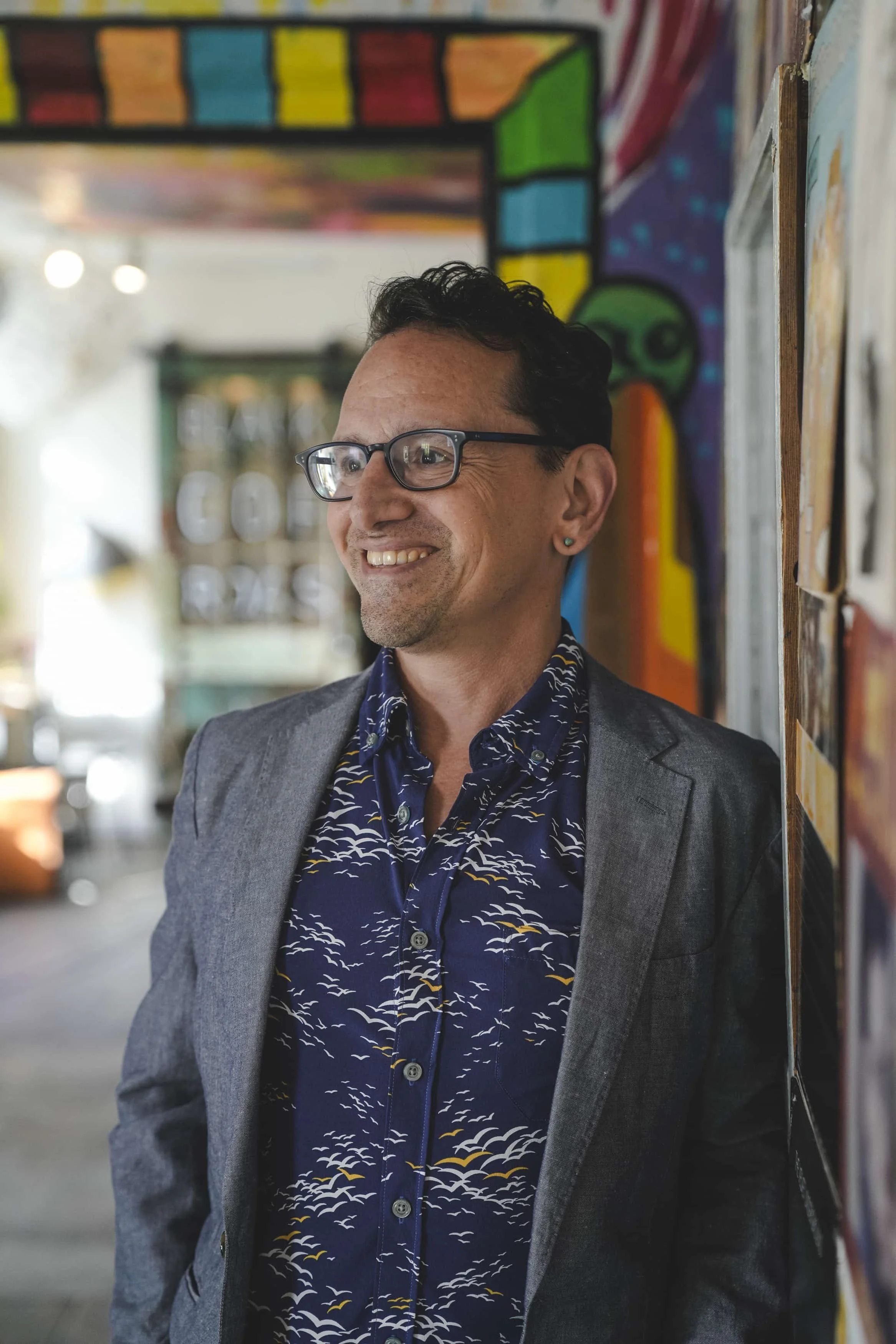 A man wearing glasses and a gray blazer, smiling, standing indoors near a colorful graffiti wall.