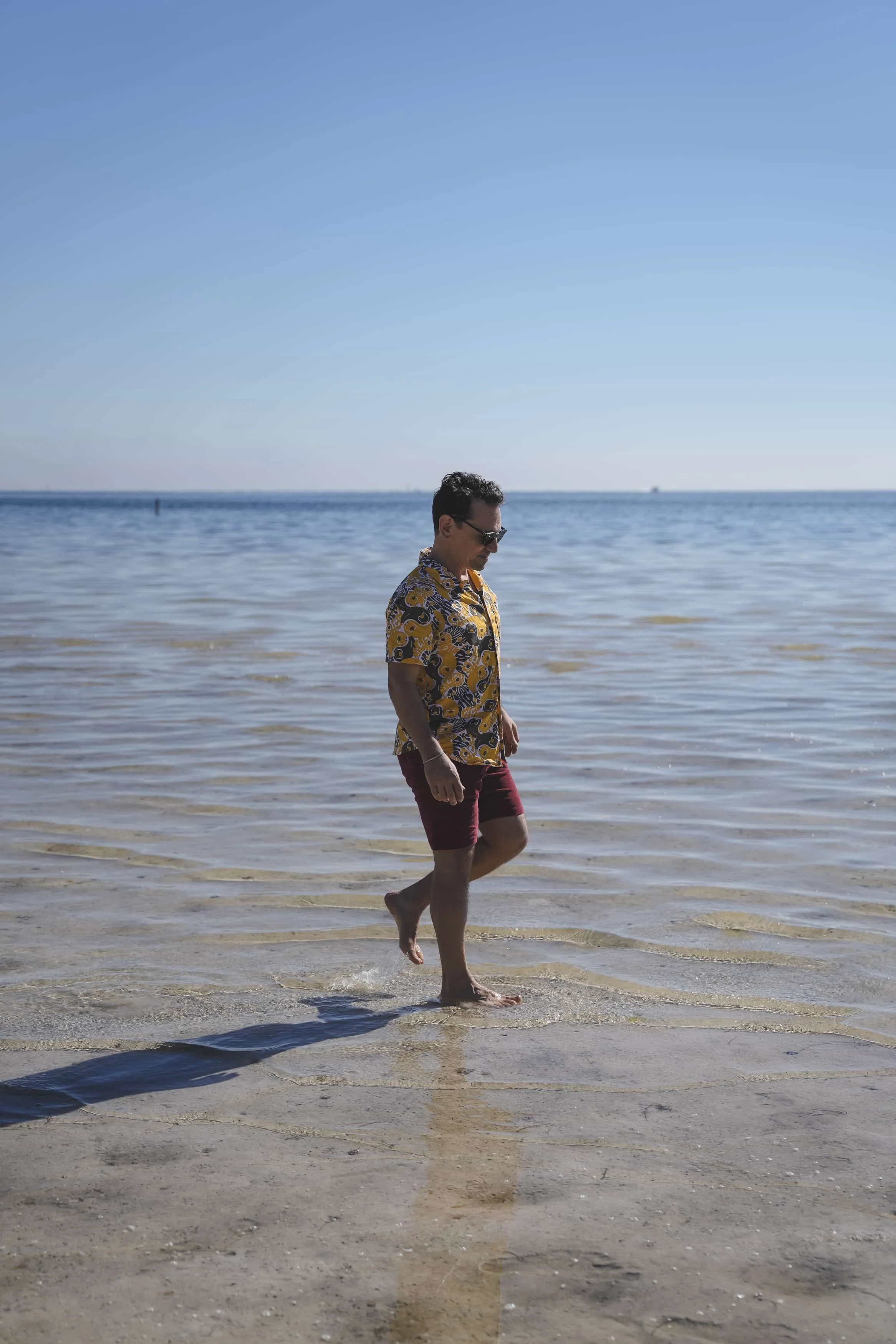 A man with dark hair, wearing sunglasses, a yellow Hawaiian shirt, and maroon shorts, walking barefoot through shallow water at the beach, with a blue sky and calm ocean in the background.