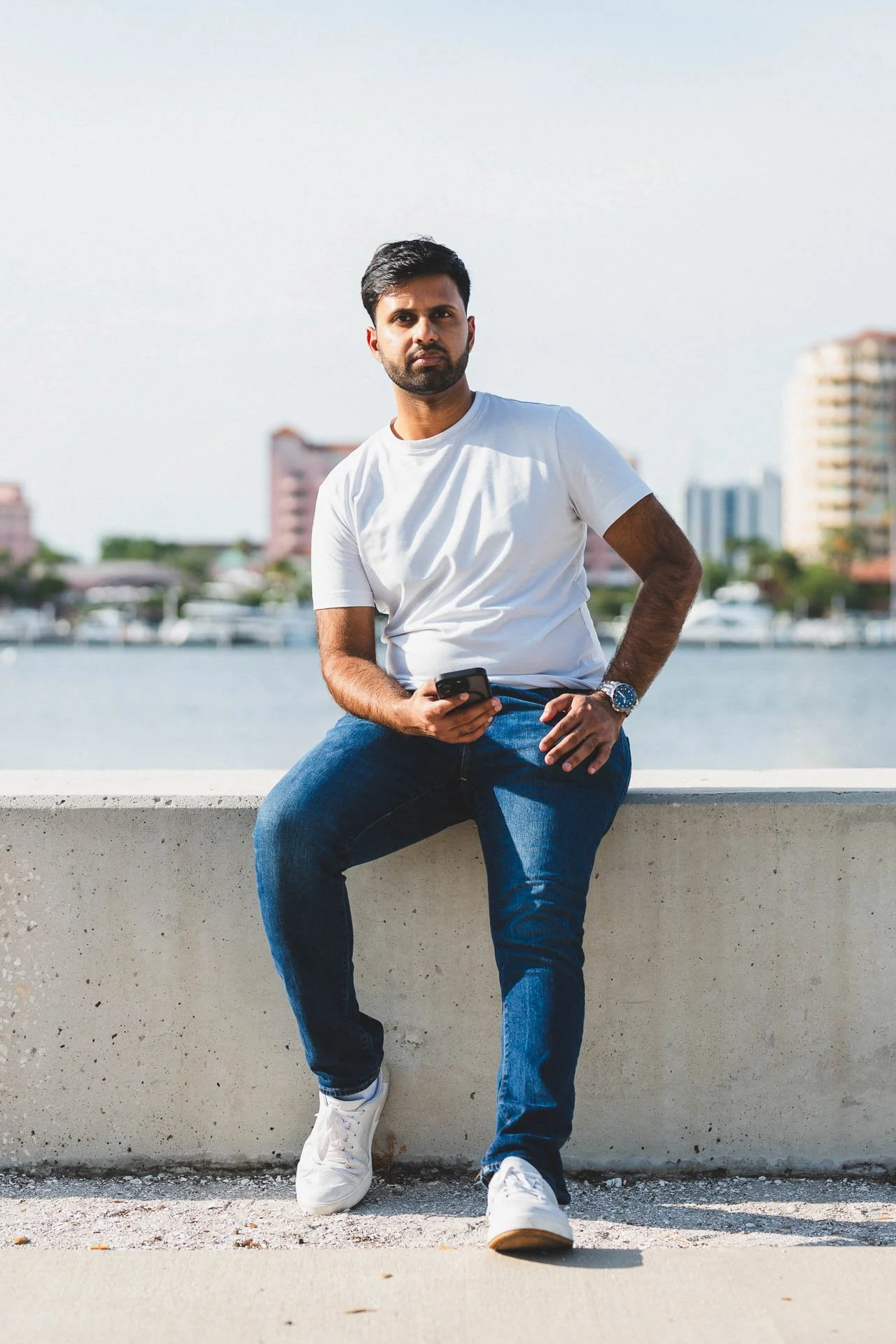 Young man with dark hair and beard sitting on a concrete ledge by a body of water, holding a smartphone, with city buildings and boats in the background on a clear day.