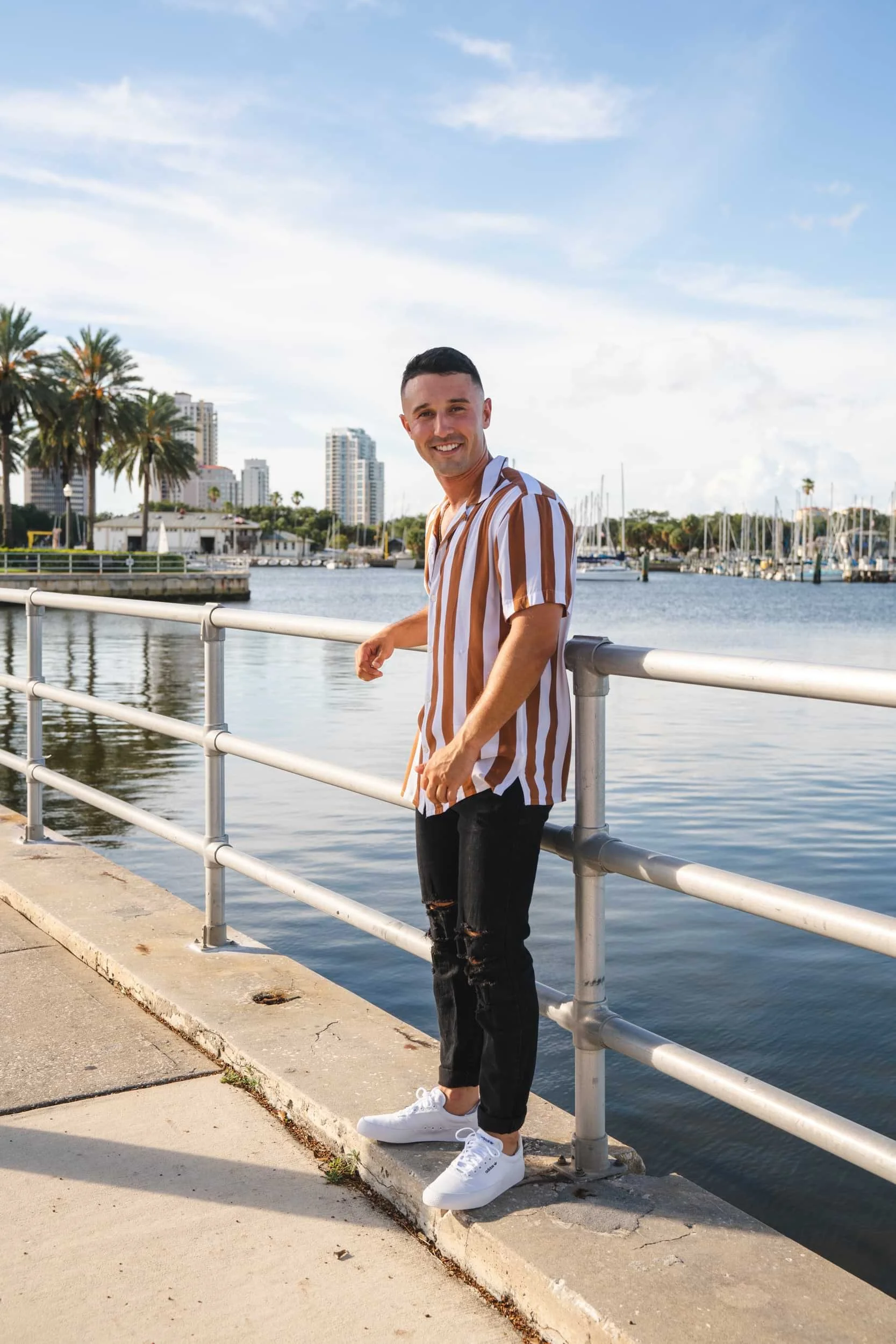 A smiling man with short dark hair, wearing a brown and white striped shirt, ripped black jeans, and white sneakers, standing by a waterfront with a city skyline and boats in the background.