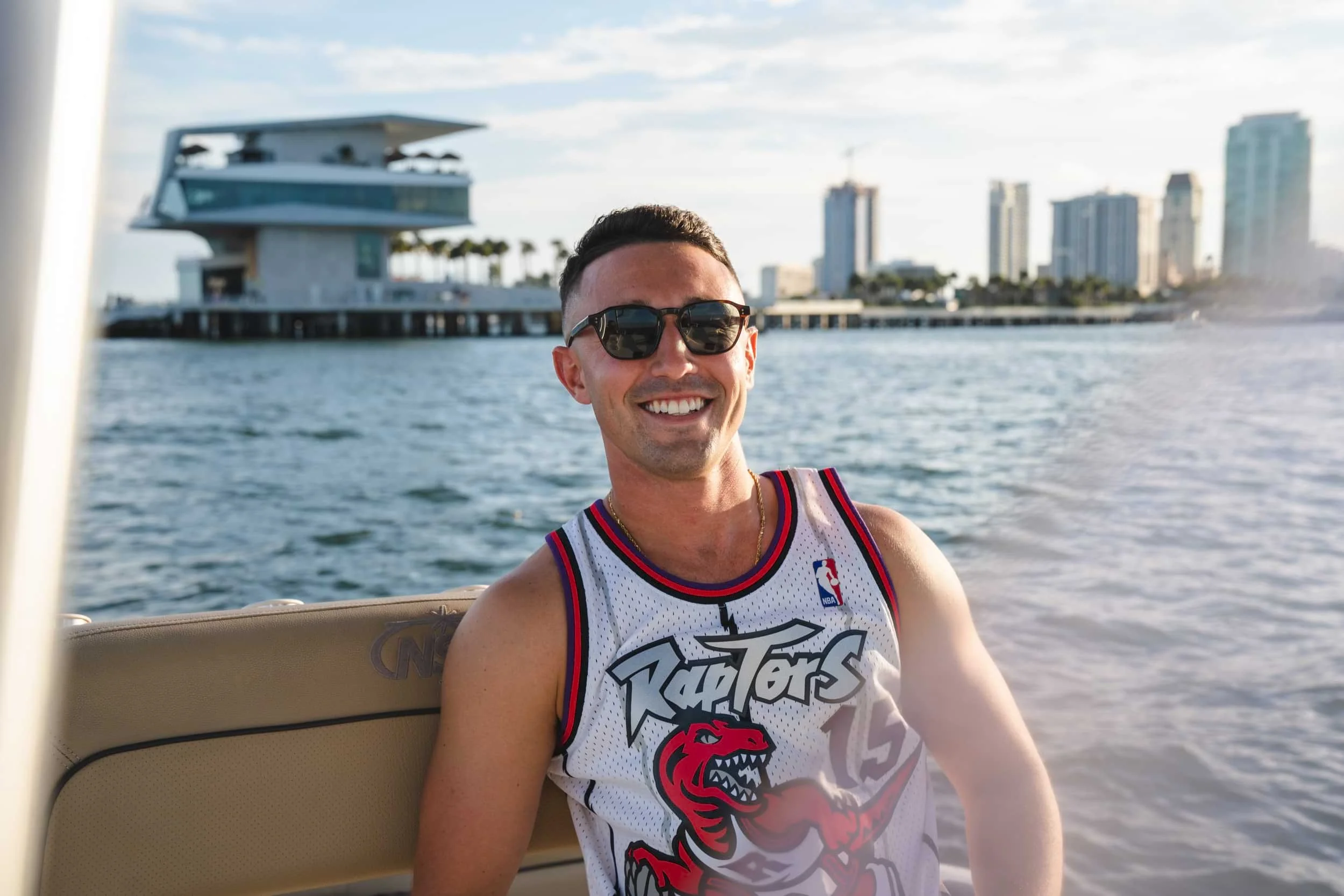 A smiling man wearing sunglasses and a white basketball jersey with a red and black dragon logo, sitting on a boat with a city skyline and water in the background.