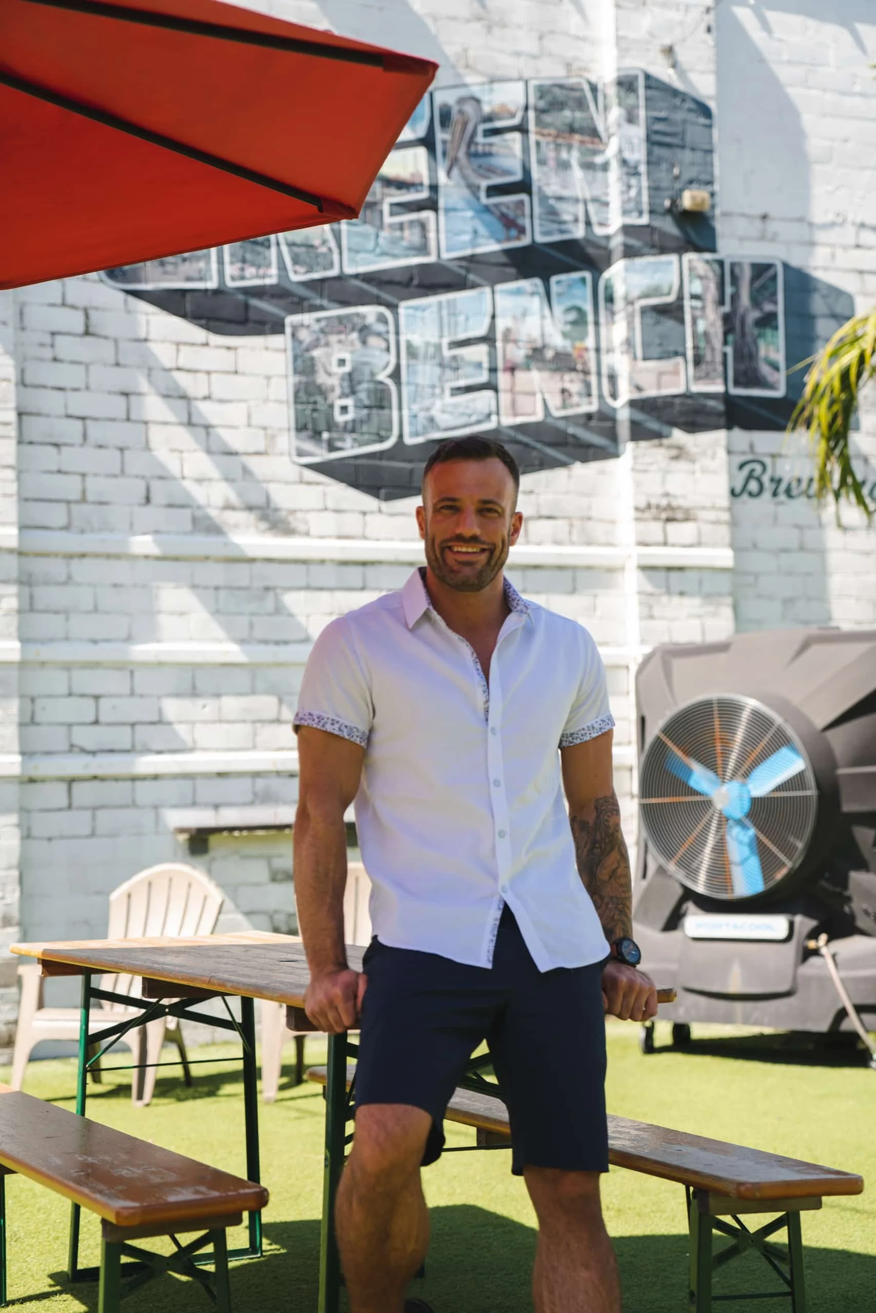 A man with short dark hair, beard, wearing a white short-sleeved button-up shirt and black shorts, standing outdoors in front of a white brick wall with a wall-mounted sign that says 'BREW'. There is a large fan on the right, a orange umbrella in the