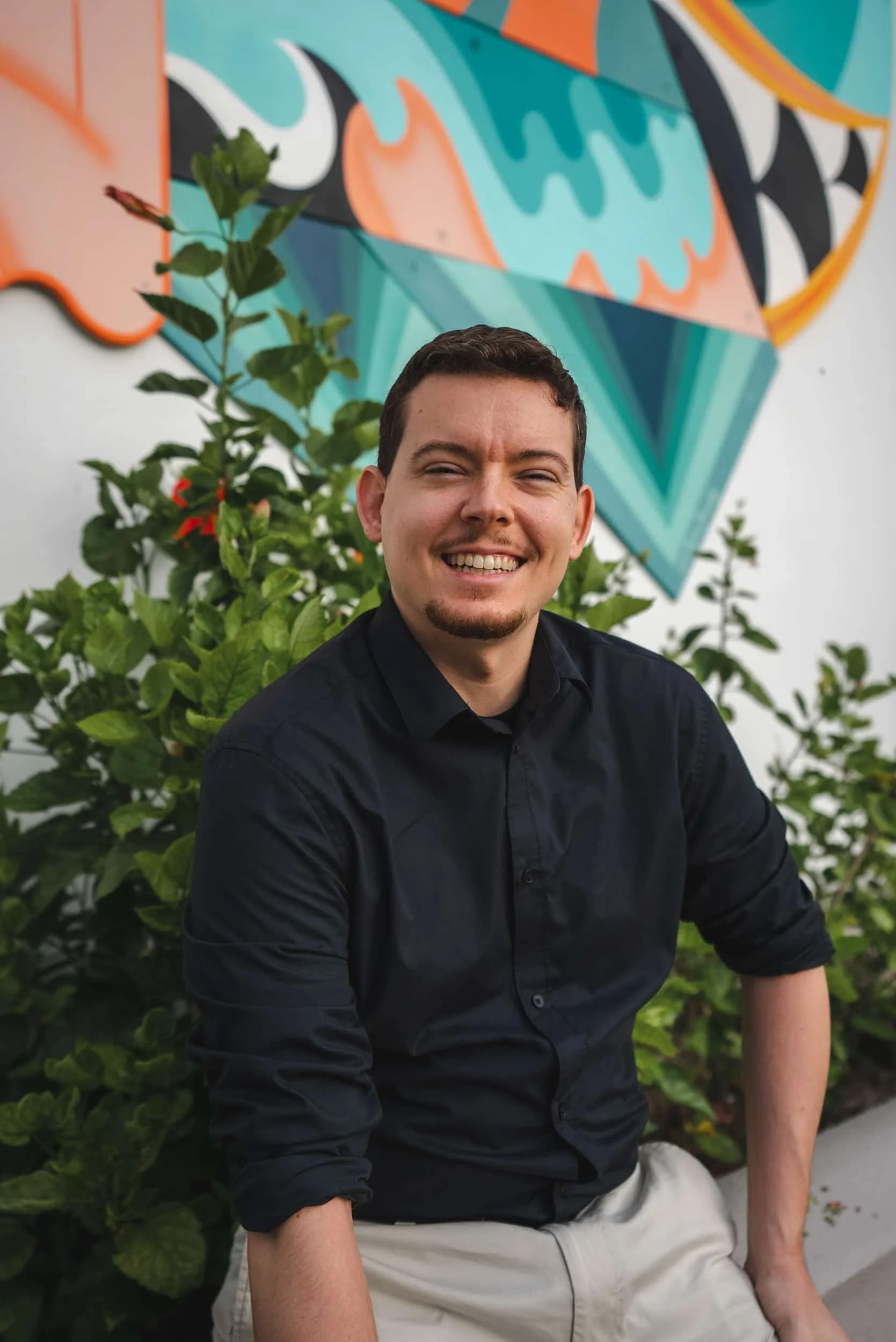 A young man smiling, sitting in front of a colorful abstract mural and green foliage.