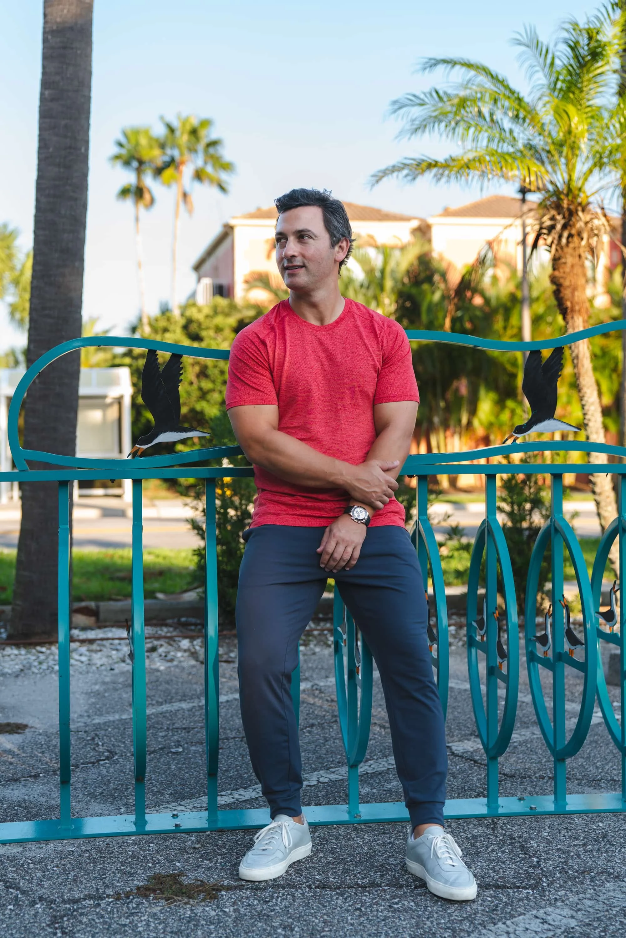 A man in a red T-shirt and gray pants leaning on a turquoise metal fence with painted penguins, outdoors with palm trees and a house in the background.