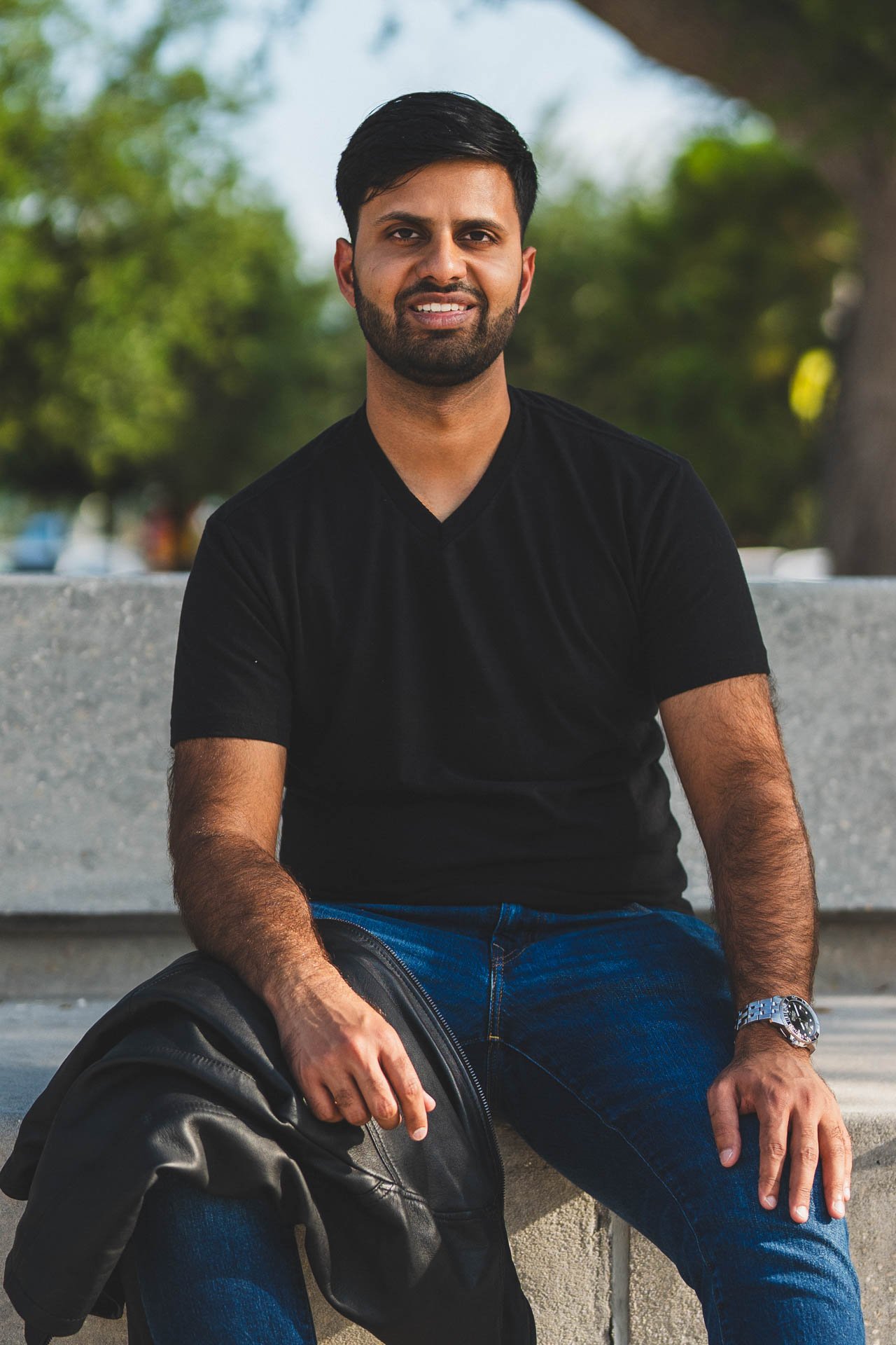 A man with dark hair and a beard sitting on a bench outdoors, wearing a black T-shirt and blue jeans, holding a black leather bag.