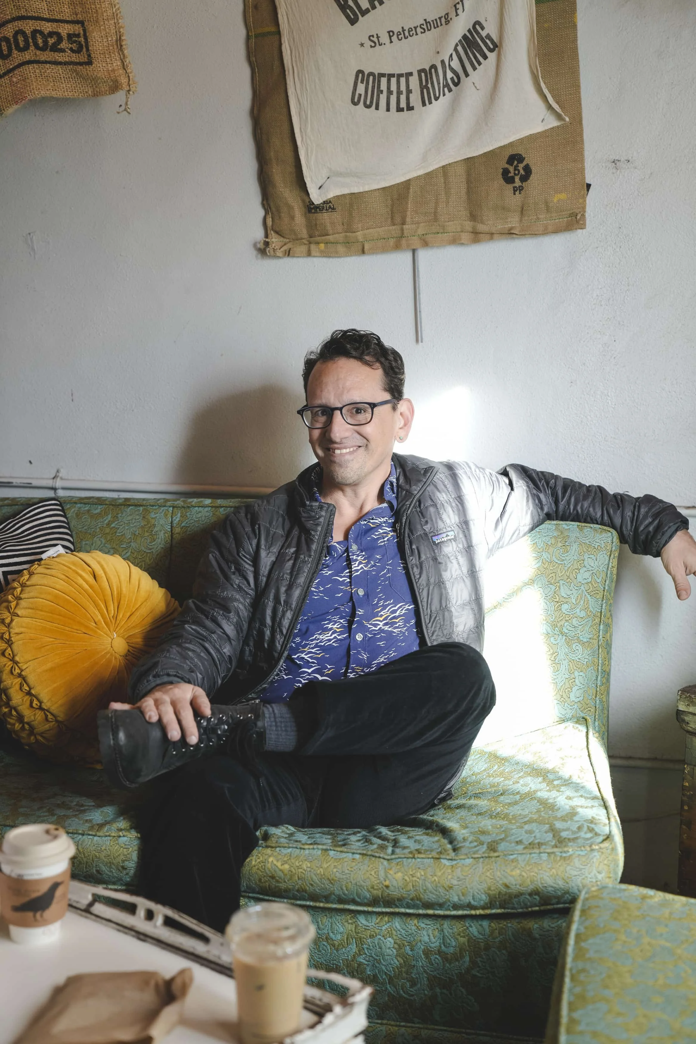 A man with glasses and dark hair, wearing a blue patterned shirt and a black jacket, sitting on an antique-style green patterned sofa, smiling at the camera, with a coffee drink on the table in front of him.