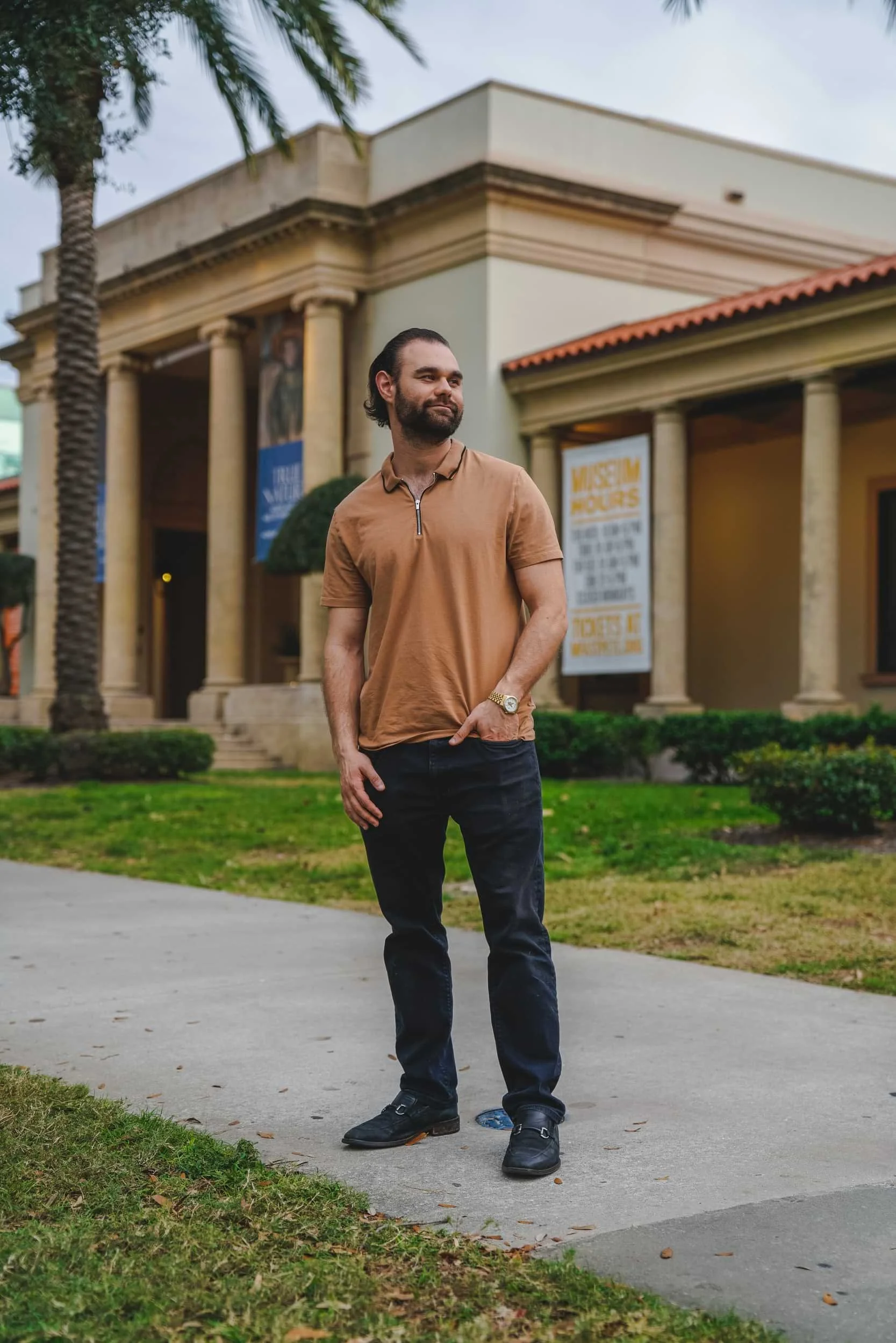 A man with a beard and long hair tied back, wearing a brown polo shirt, dark jeans, and black shoes, standing on a sidewalk with grass and trees, in front of a building with columns and posters, during daytime.