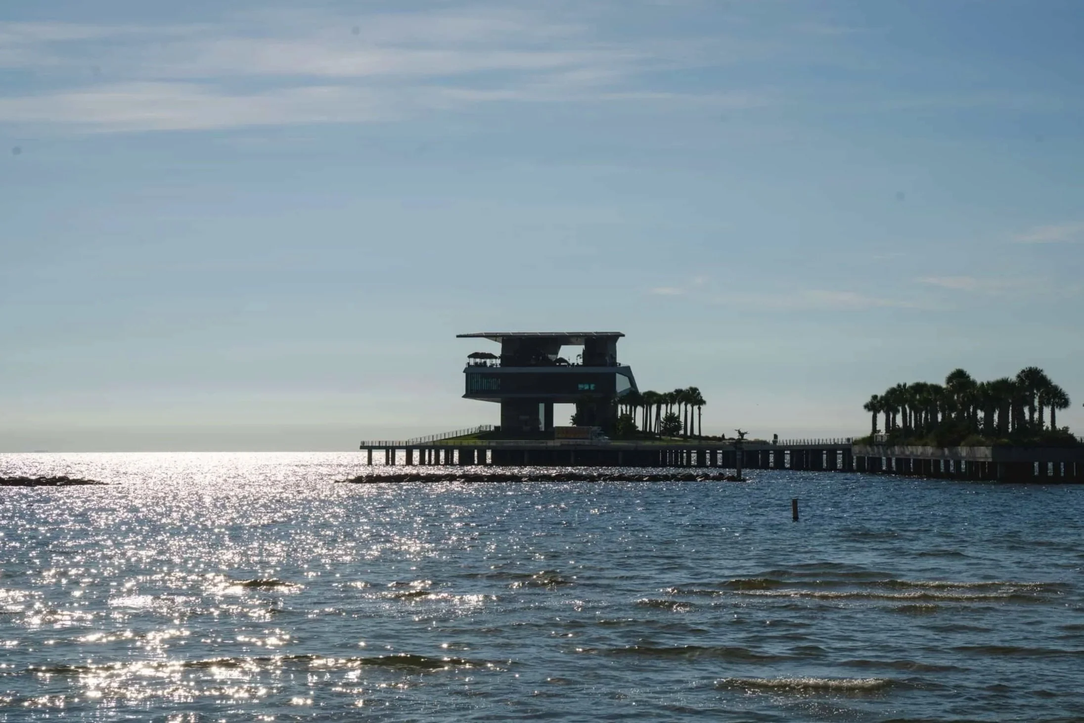 A pier extending into the ocean with a modern two-story building on stilts at the end, surrounded by palm trees, under a partly cloudy sky with sunlight reflecting off the water.