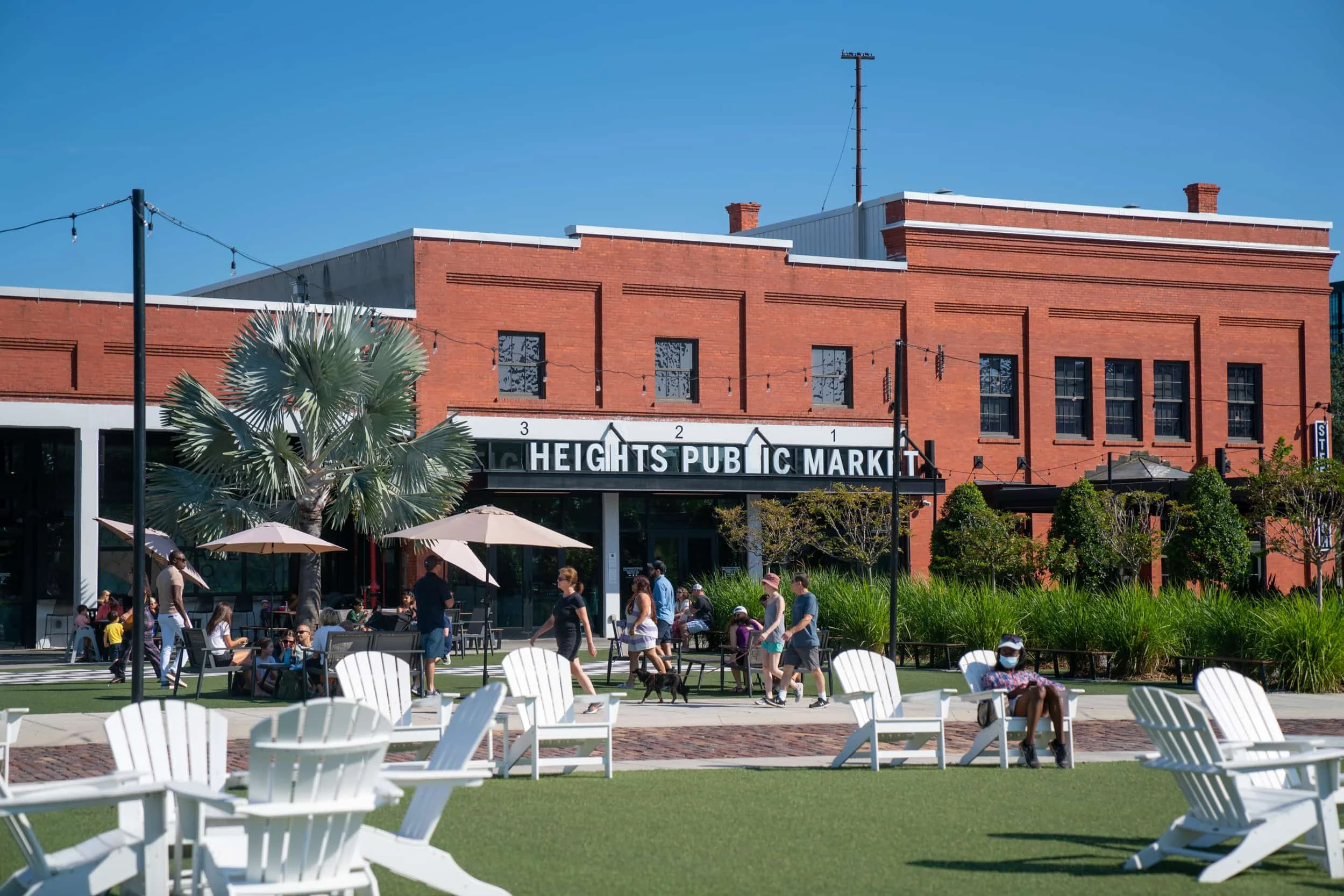 People enjoying outdoor seating at Heights Public Market with white chairs, umbrellas, and greenery in front of a brick building under a clear blue sky.