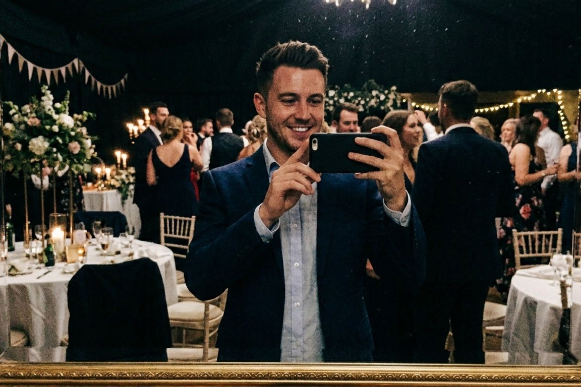 A man in a suit taking a selfie at a wedding reception with many guests in the background, decorated with flowers, candles, and fairy lights.