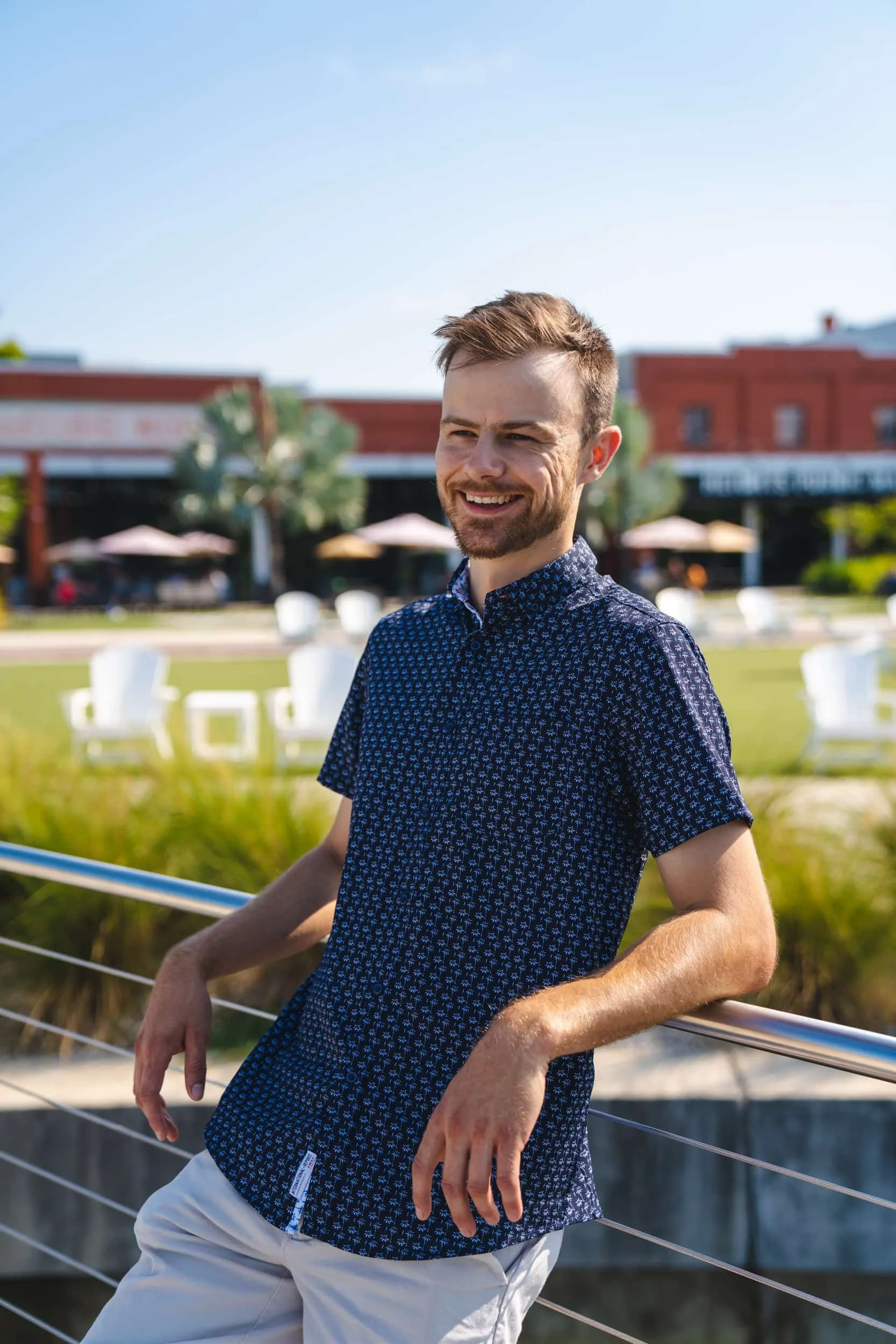 A young man with short brown hair and a beard smiling outdoors on a sunny day, wearing a blue patterned shirt and white pants, leaning on a railing with a background of a park or outdoor dining area with umbrellas and buildings.