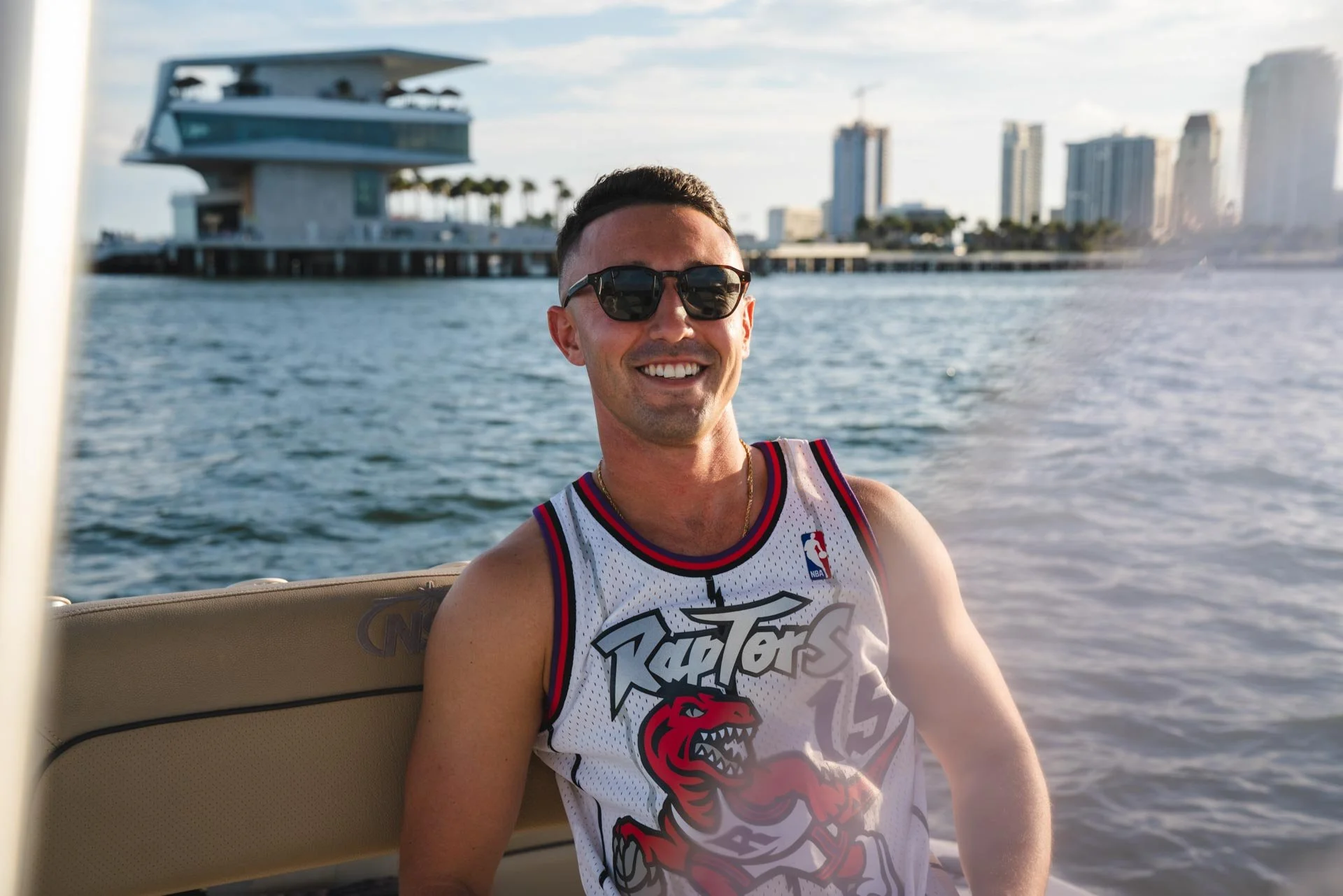 A smiling man wearing sunglasses and a white basketball jersey with a red dinosaur logo on a boat on the water with a city skyline in the background.