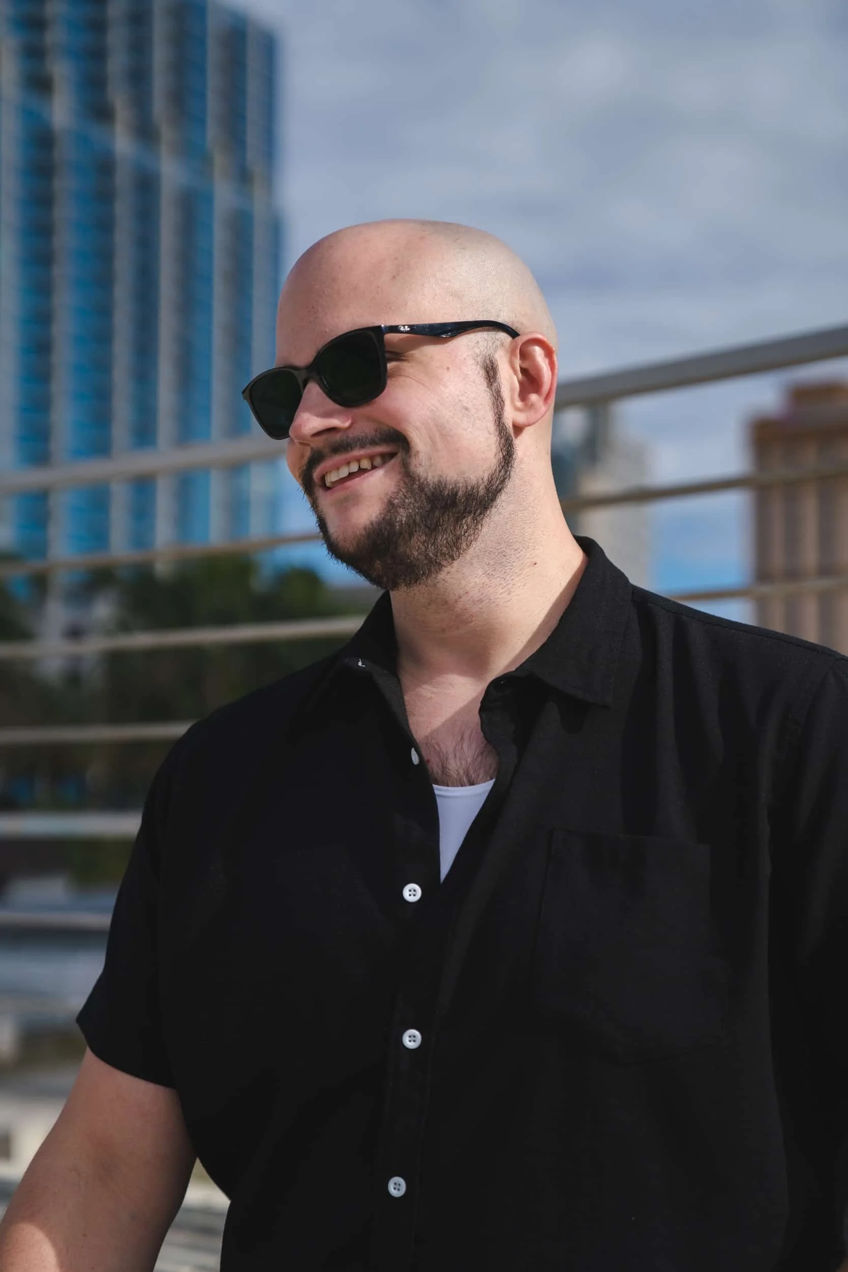 A smiling man with a beard wearing black sunglasses and a black shirt, outdoors with buildings and a blue sky in the background.